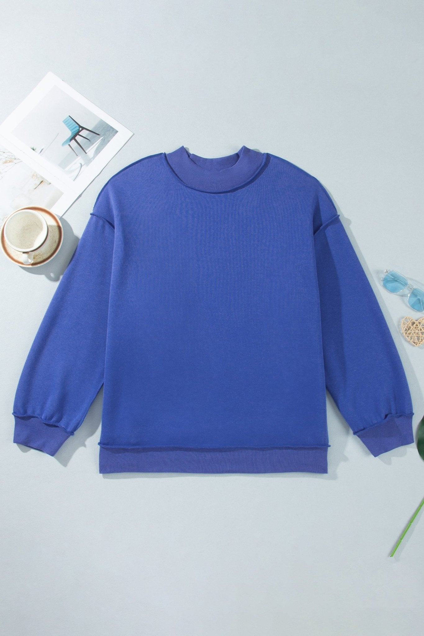 Royal blue long-sleeve crewneck sweater laid flat on light gray with cup, card, and decorative elements.