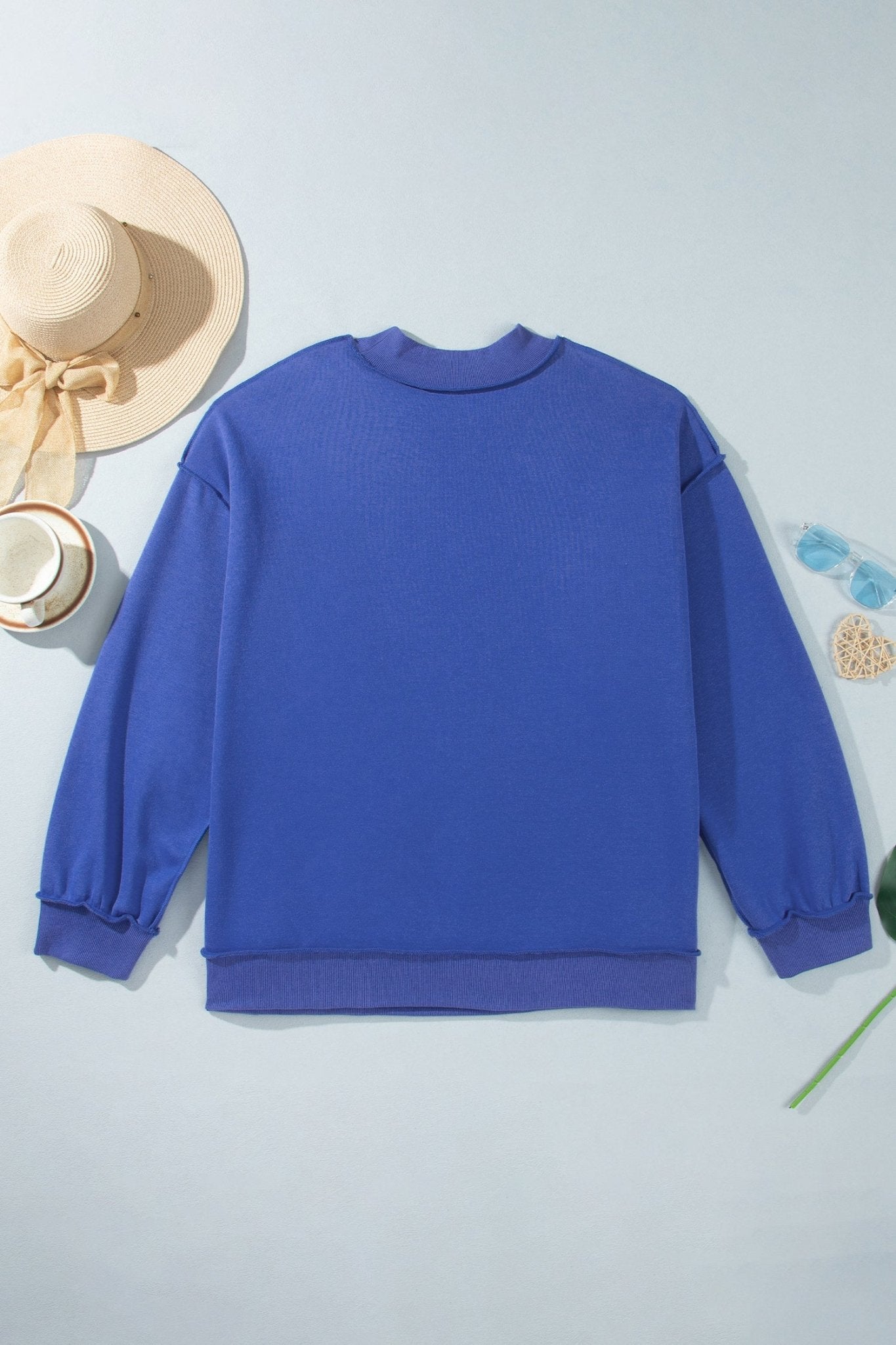 Royal blue crewneck sweatshirt laid flat on light blue with straw hat, cup, and sunglasses.