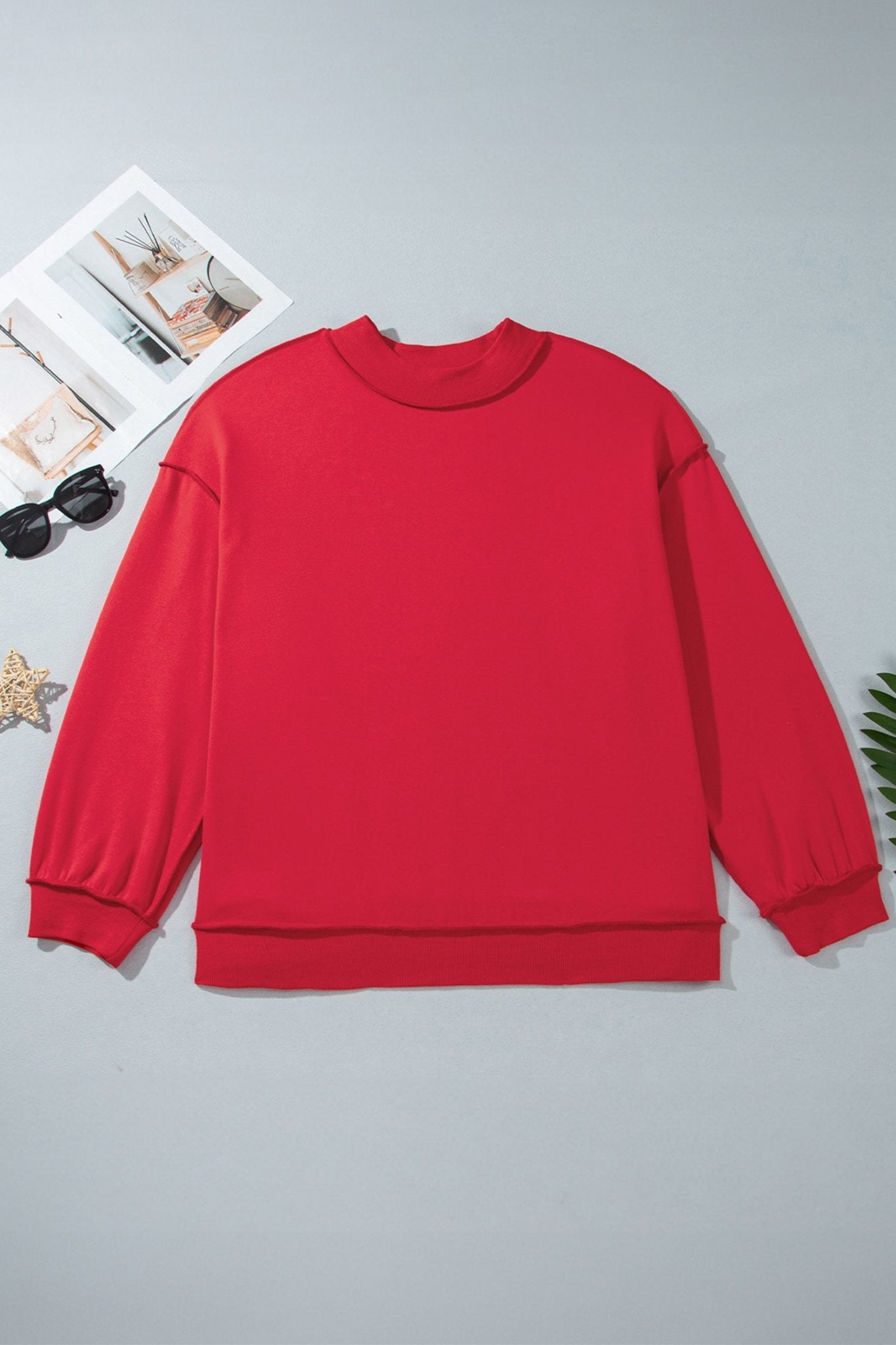 Bright red crewneck sweatshirt laid flat on light gray with sunglasses, magazine page, and star.