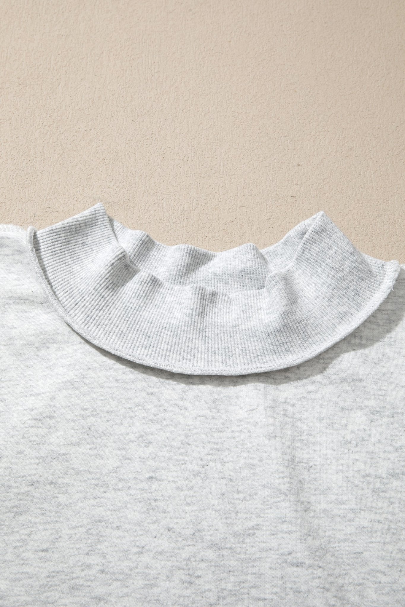 Close-up of a light gray heather knit top with a wide, ruffled Peter Pan collar against a beige textured background.