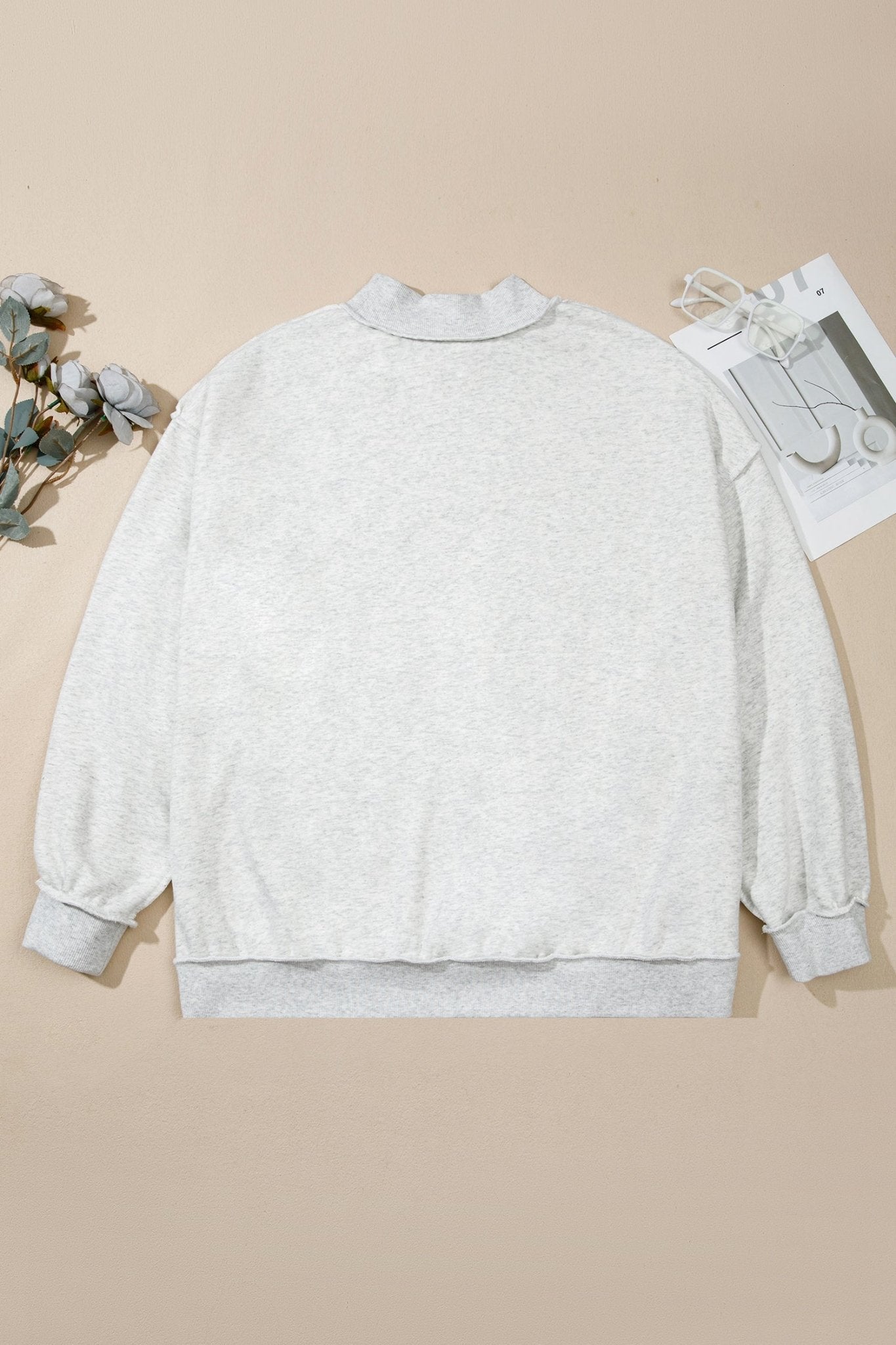 Light gray heather crewneck sweatshirt laid flat on beige with dried flowers and a magazine page.