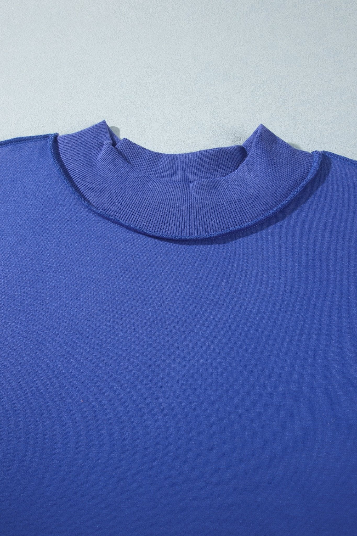 A close-up of a royal blue knit top showing the ribbed mock neck collar against a light gray background.