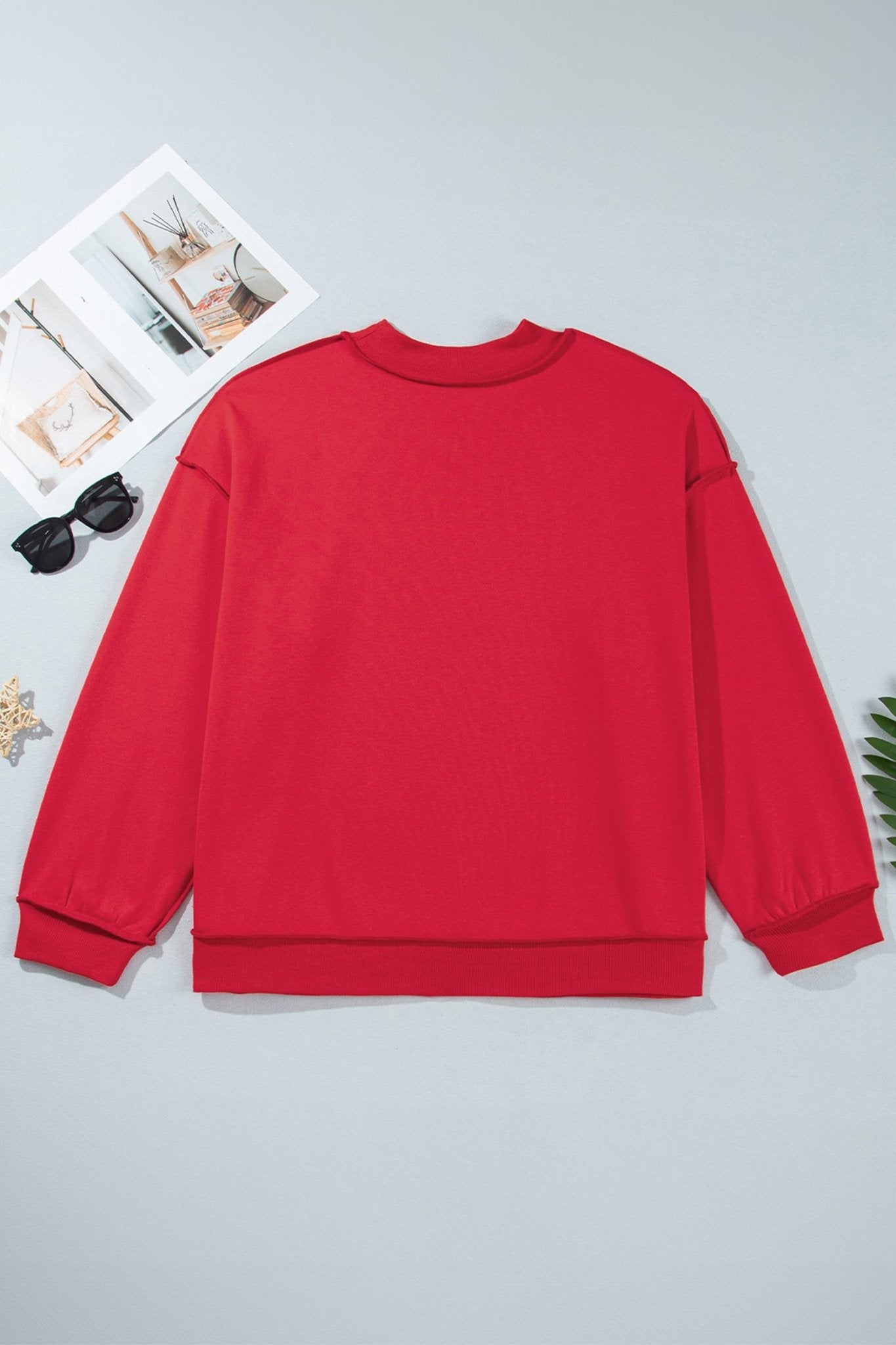 Bright red long-sleeve crewneck sweatshirt laid flat on light gray with sunglasses, magazine, and leaf.
