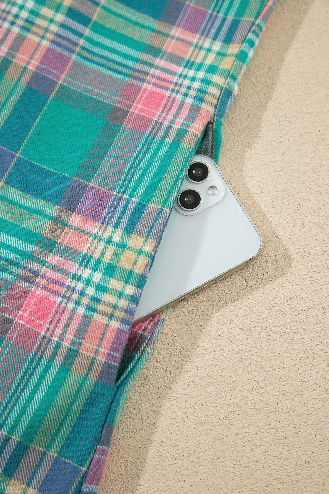 Light blue smartphone peeking from teal, pink, and green plaid fabric on textured beige.