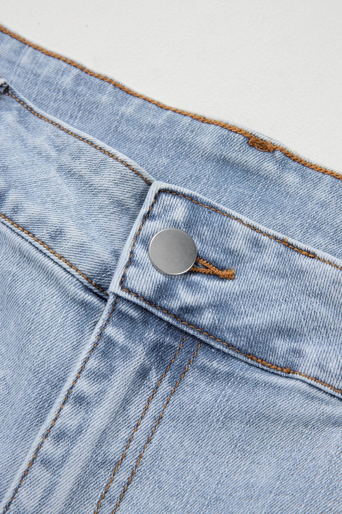 Close-up detail of a pair of light blue denim jeans showing the button closure, metal button, and contrast stitching.