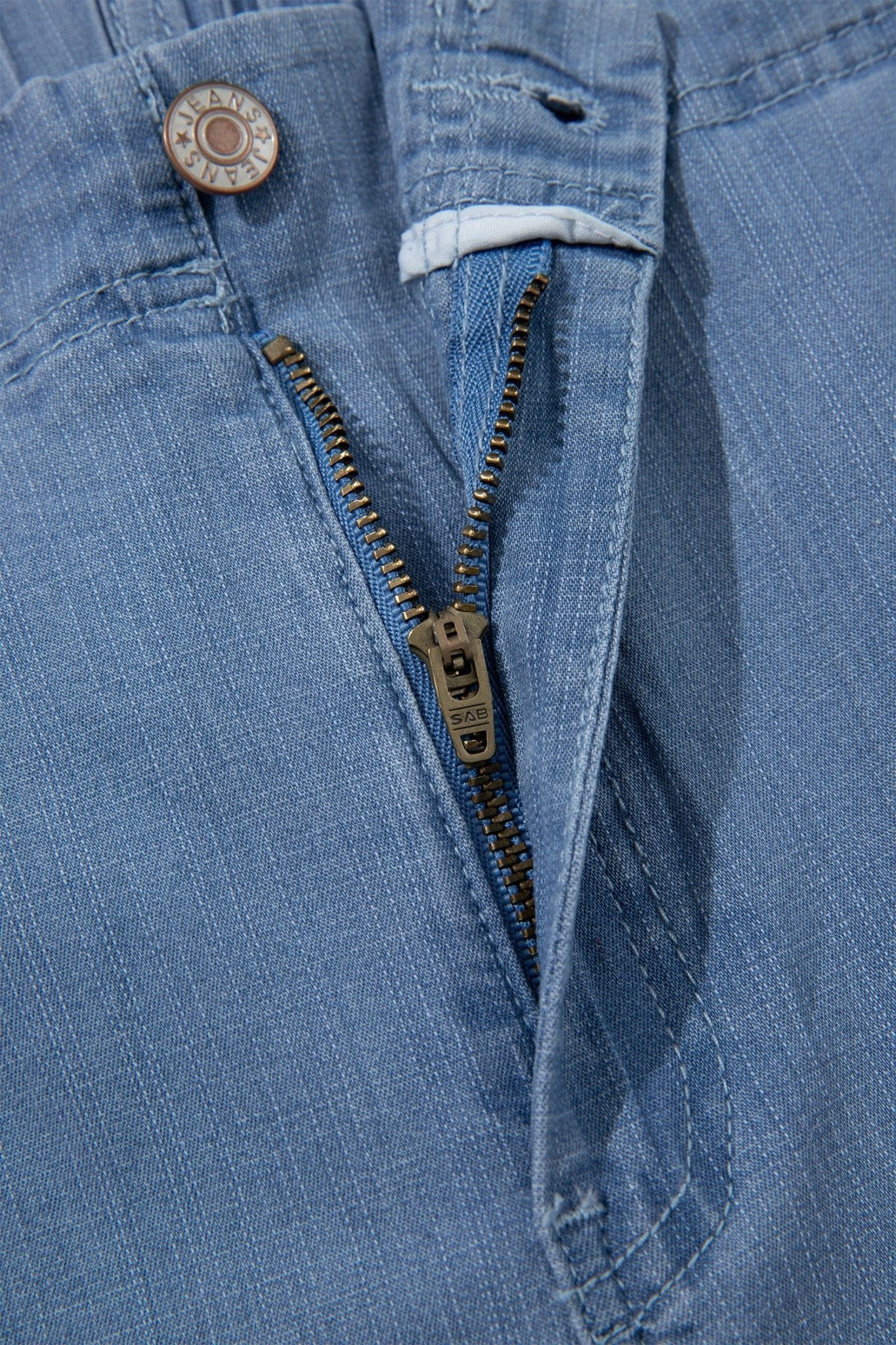 Close-up of a light blue denim garment, showing the waistband, button, zipper, and fabric texture.