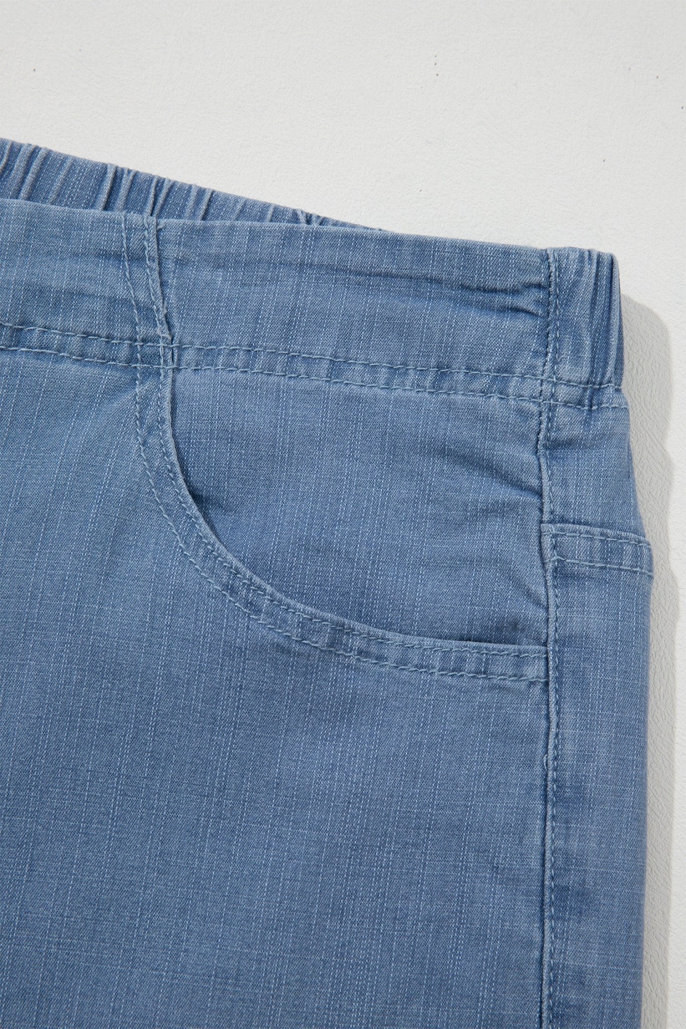 Close-up of blue denim pants showing the waistband and front pocket seam against a light background.