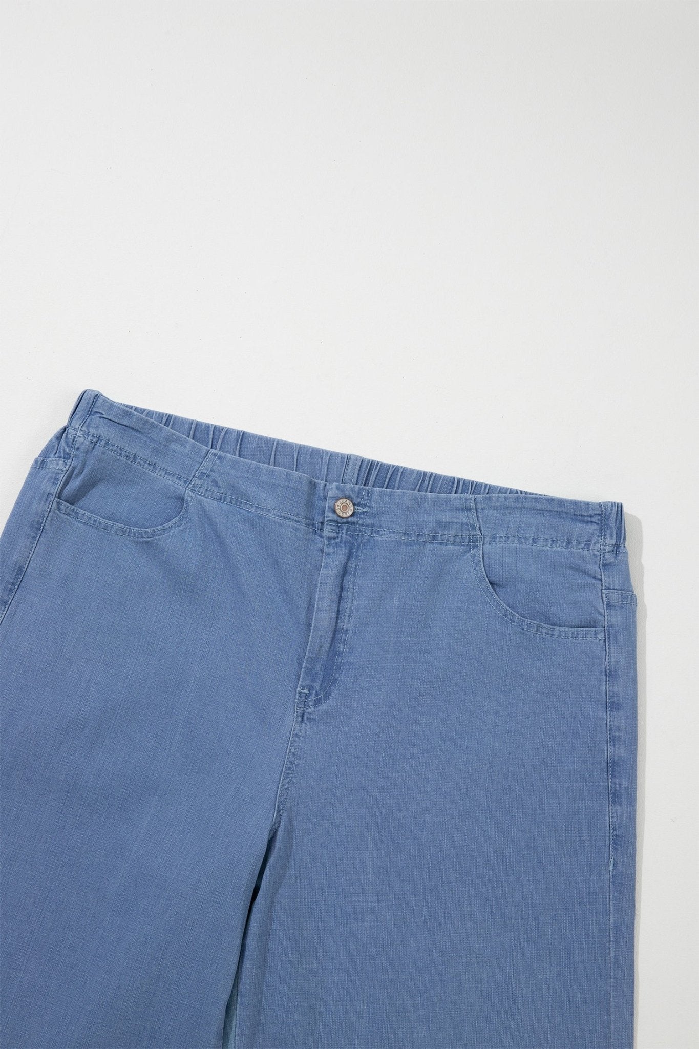 Close-up of light blue denim jeans showing waistband, button closure, belt loops, and front pockets on light gray.