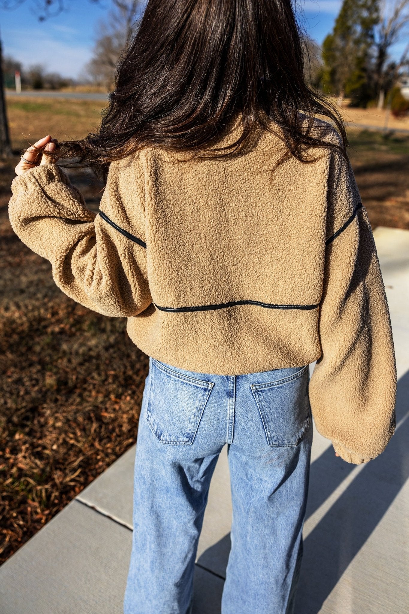Back view of person in tan sherpa sweater with black piping and light jeans.