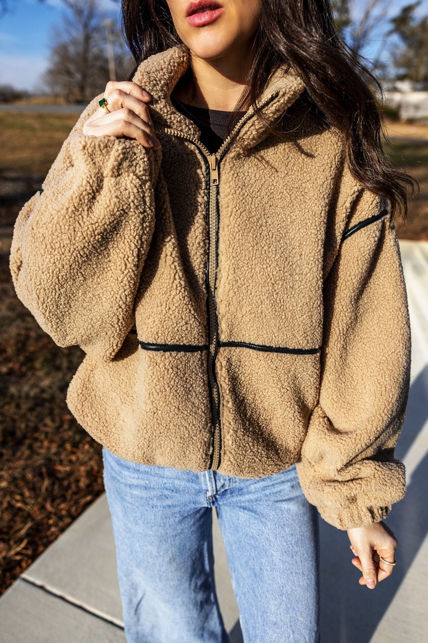 Woman in tan sherpa fleece jacket with black trim and light blue jeans.