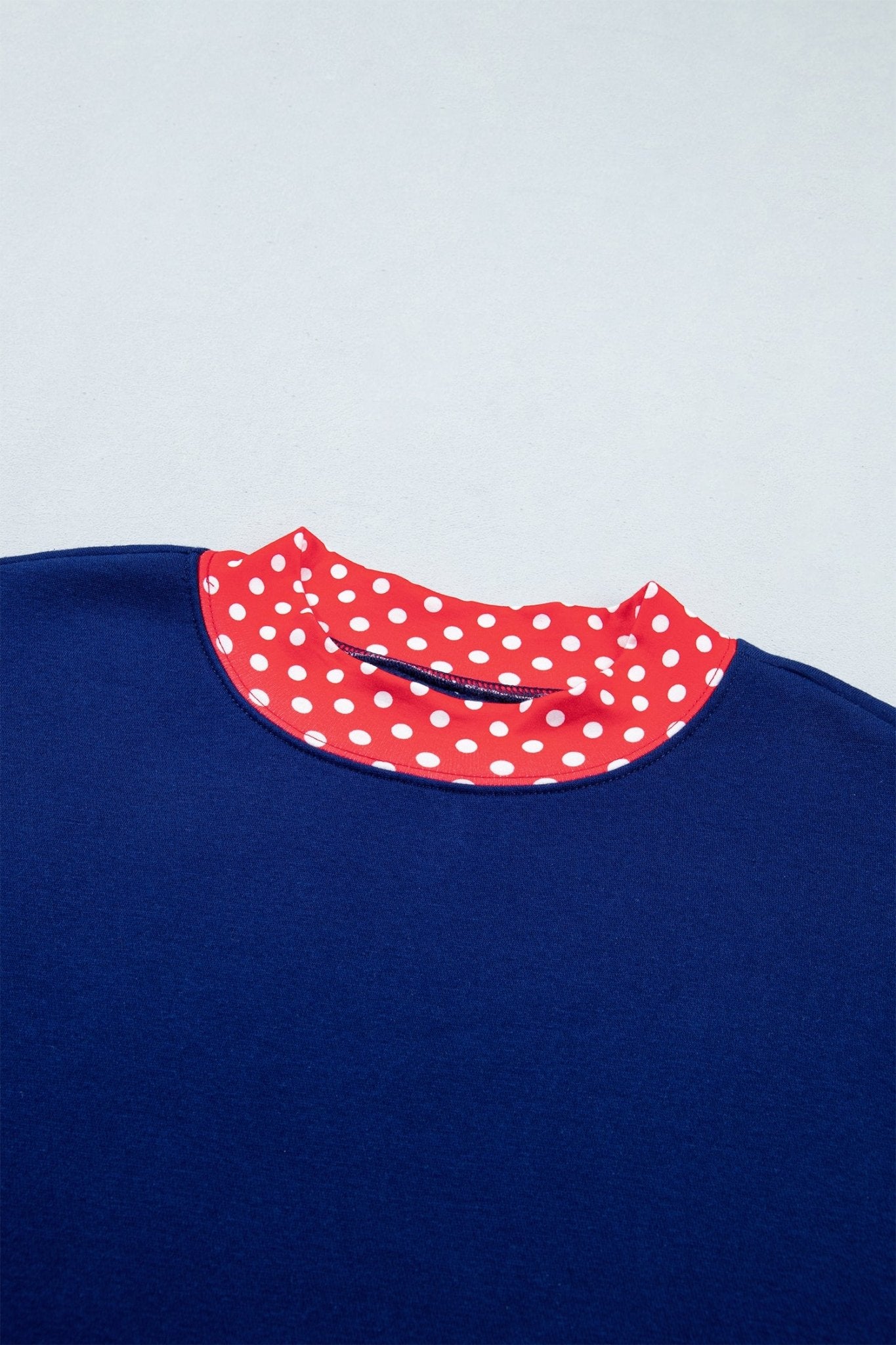 Close-up of navy blue crewneck sweater with red collar featuring white polka dots on light gray background.