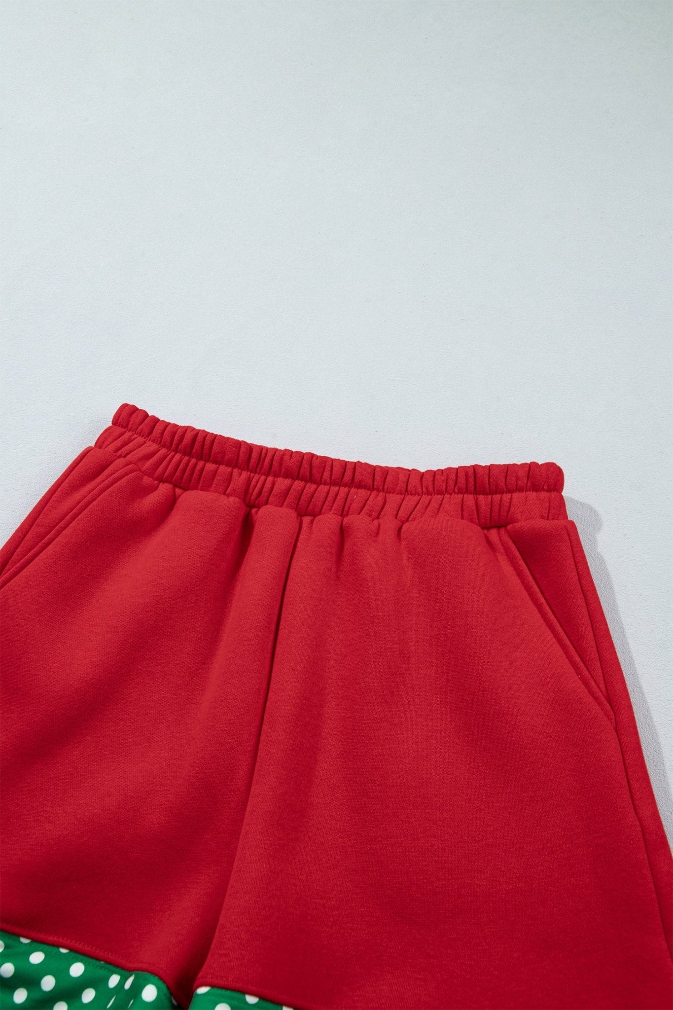 Close-up of bright red shorts with elastic waistband and green-white polka-dot trim on light gray.