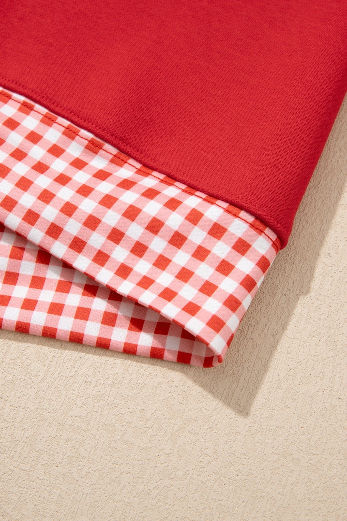 Folded red sweatshirt with red-white gingham check reverse side on a textured beige surface.