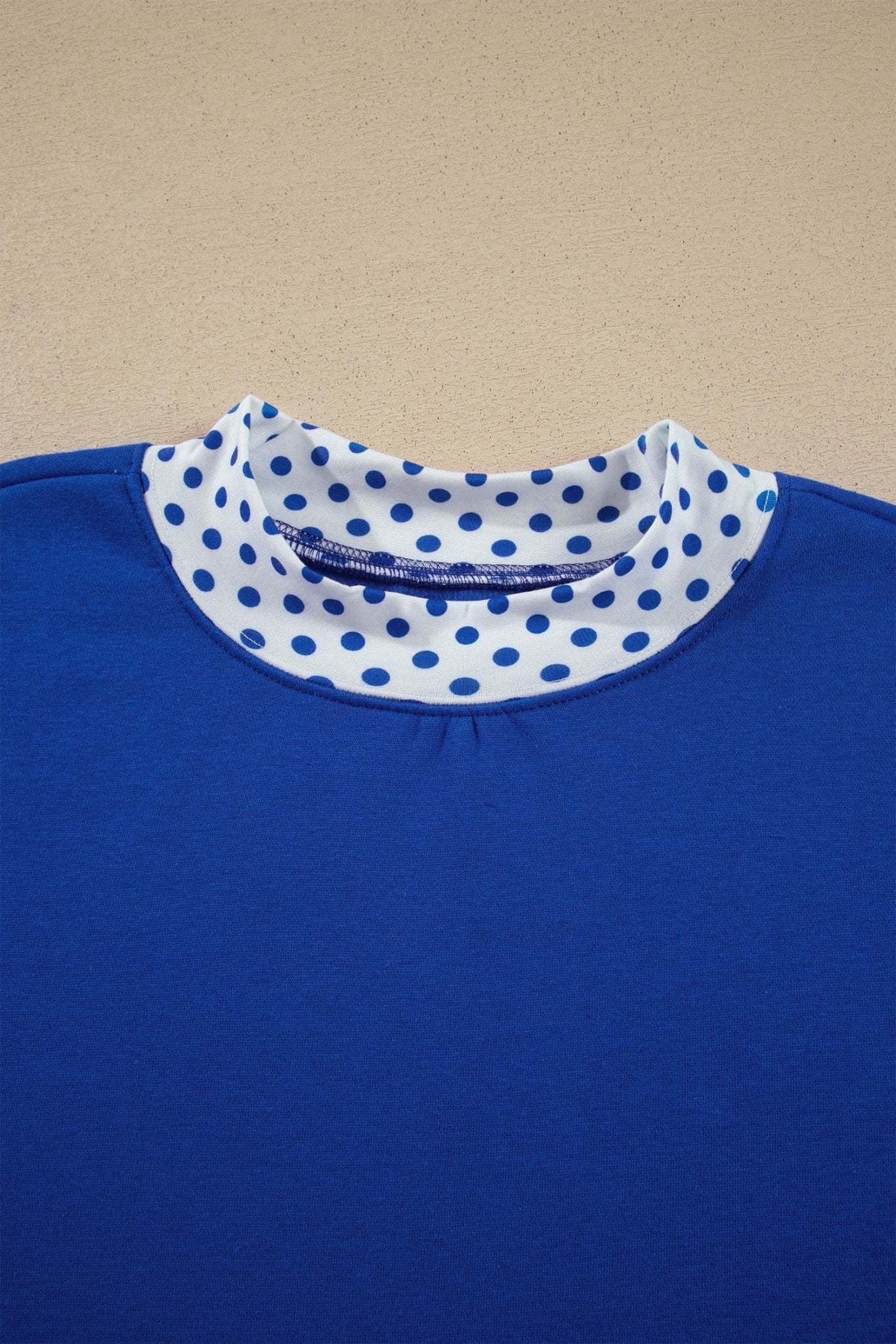 Close-up of royal blue top with contrasting white mock neck collar featuring blue polka dots against beige background.