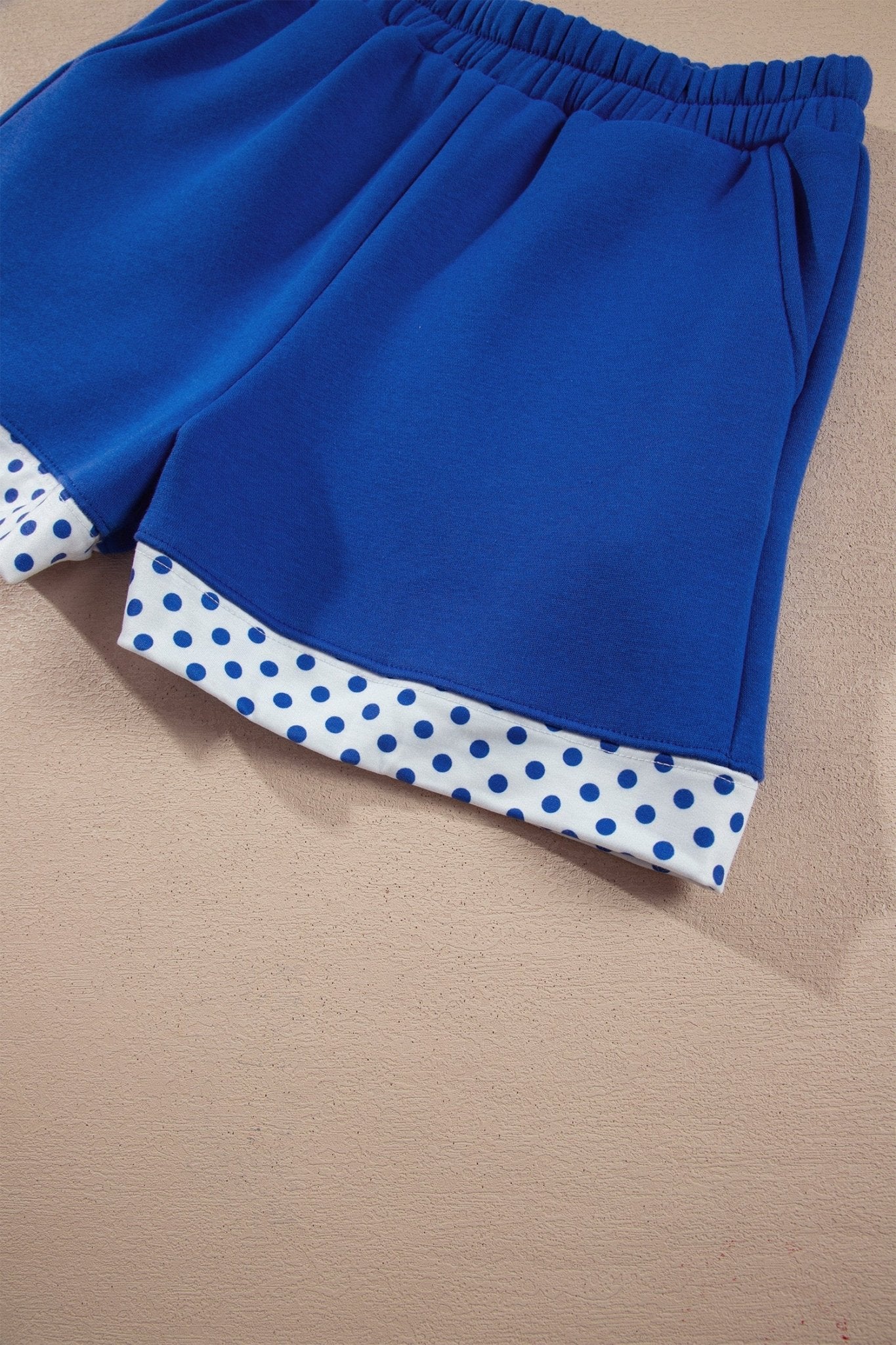 Blue shorts with a white and blue polka dot hem, shown on a beige textured background.