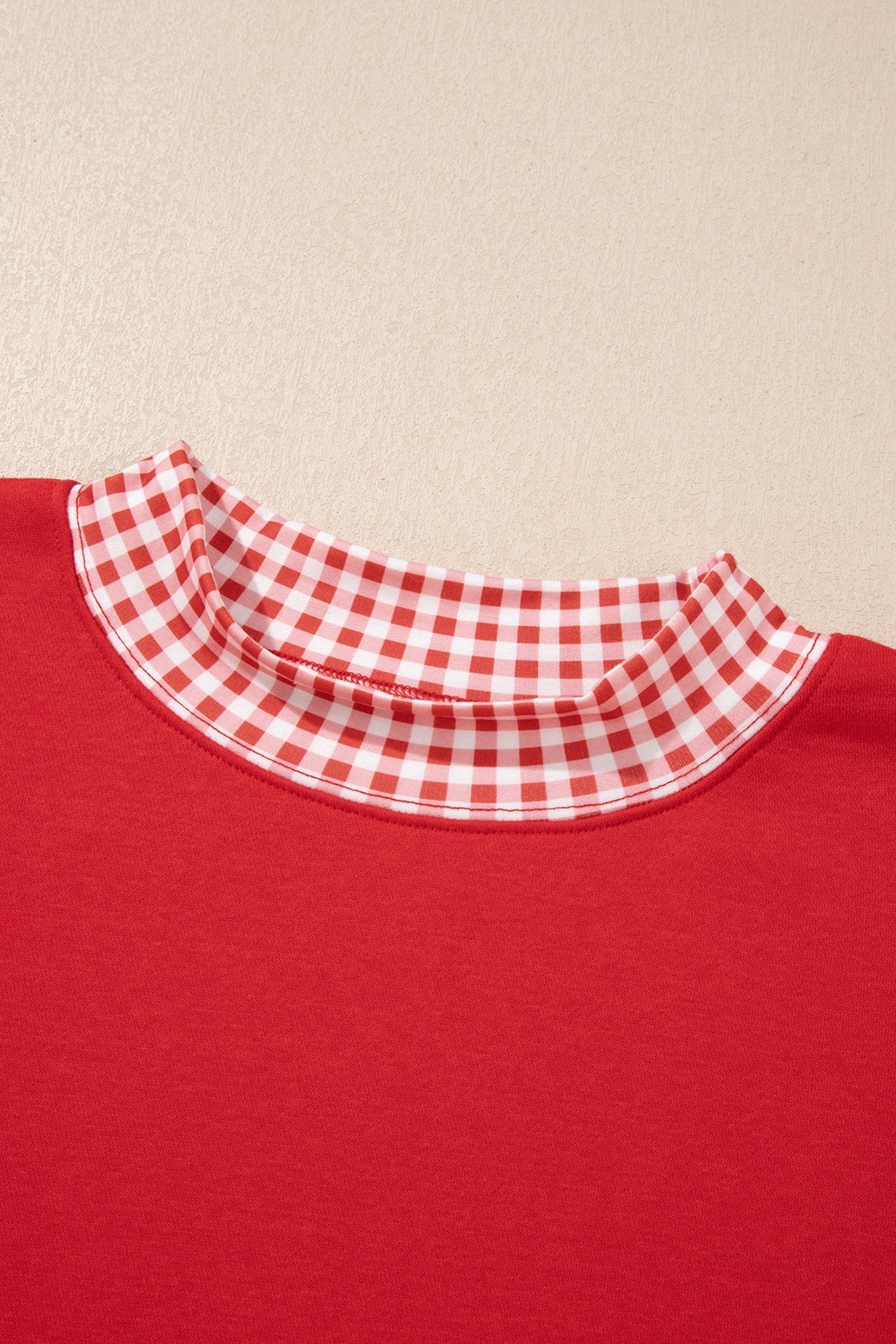 Close-up of red crewneck shirt with contrasting red-white gingham check collar against a beige background.