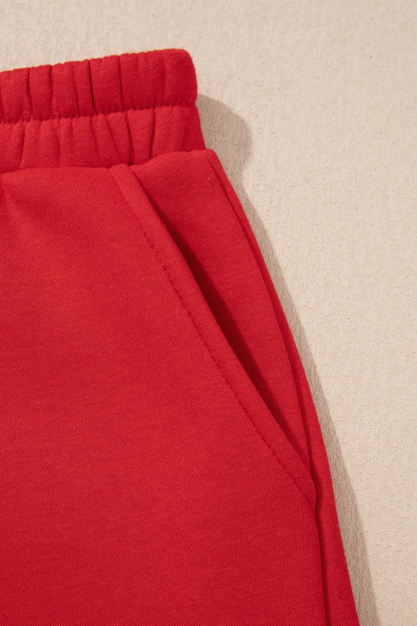 Close-up of bright red sweatpants with elastic waistband and side pocket seam on a beige background.