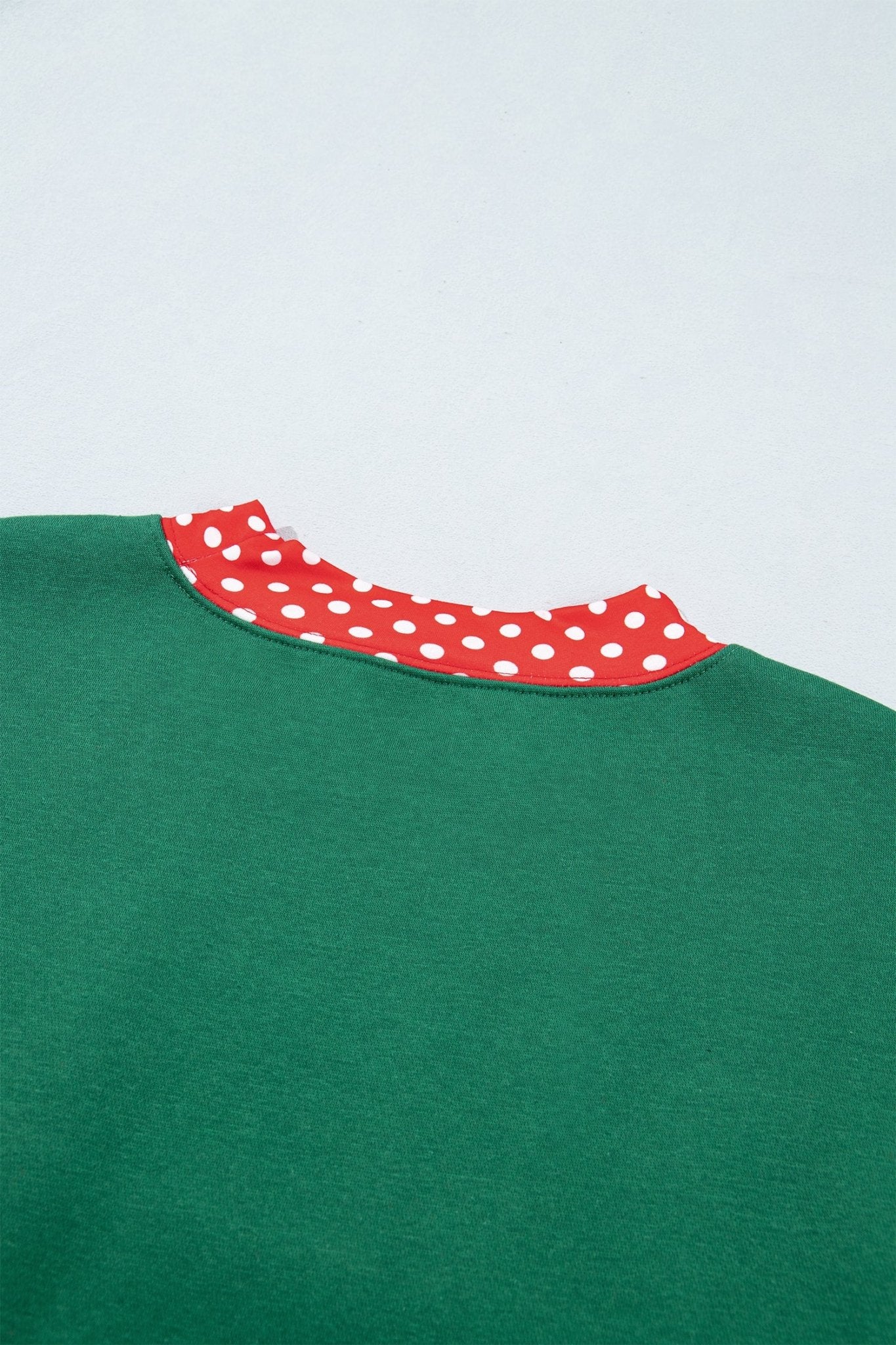 Close-up of green crewneck sweatshirt with contrasting red collar featuring white polka dots on a light gray background.