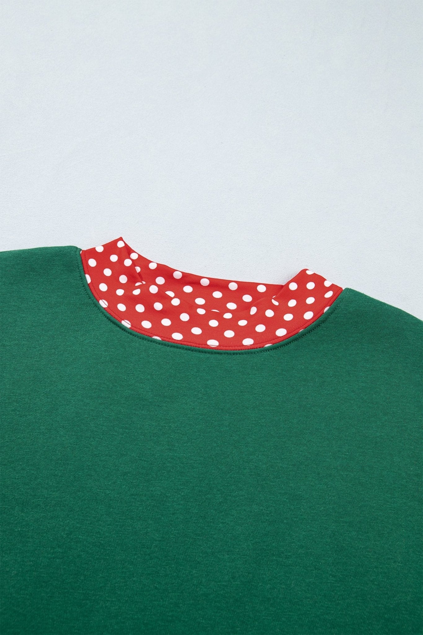 Close-up of green crewneck sweater with red collar featuring white polka dots against light gray background.
