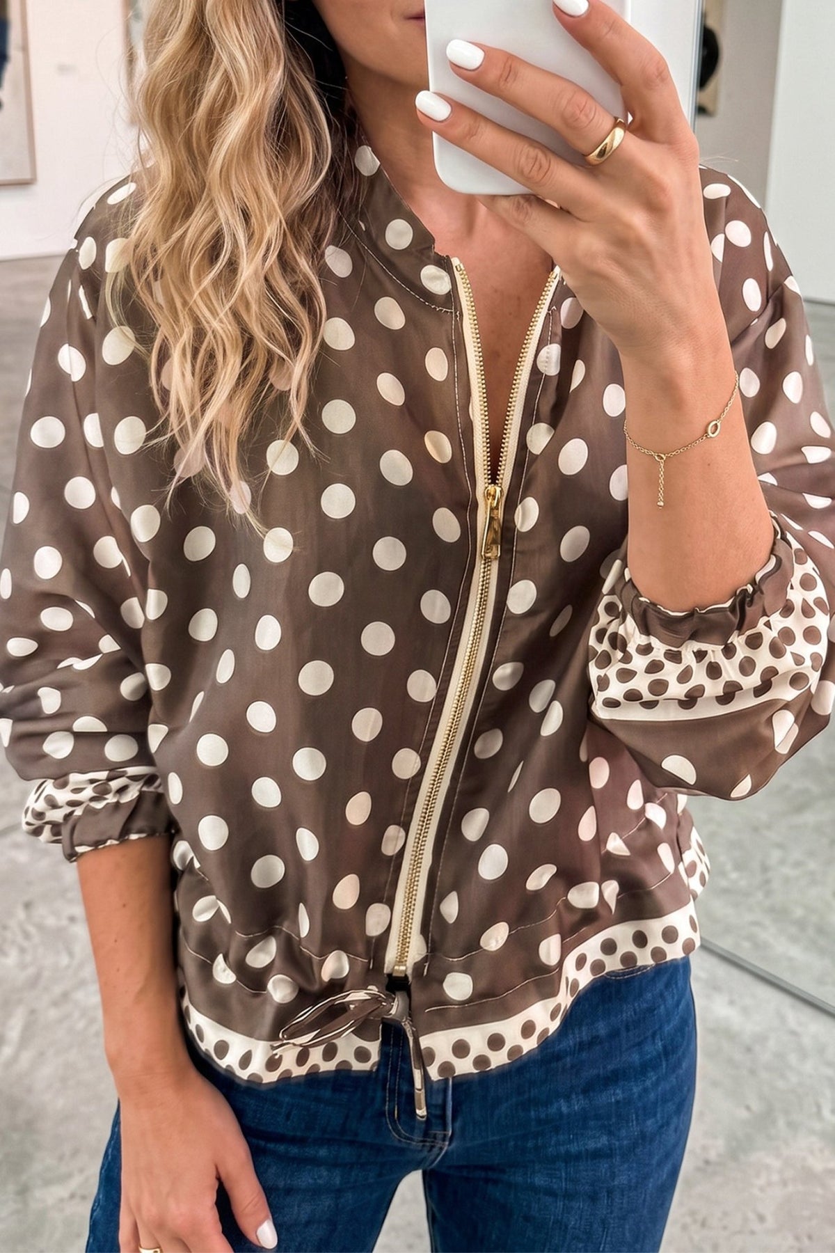 Woman in brown and beige polka dot zip-up jacket with ruffled trim and blue jeans, mirror selfie.