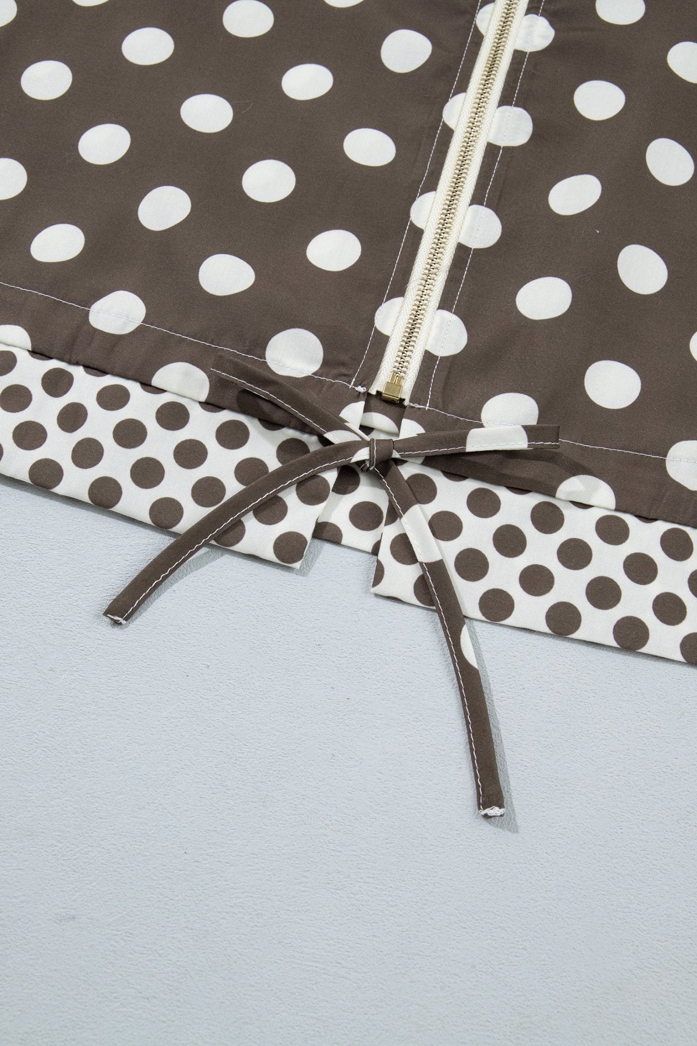 Close-up of a brown and white polka dot fabric item with a cream zipper and matching tie detail