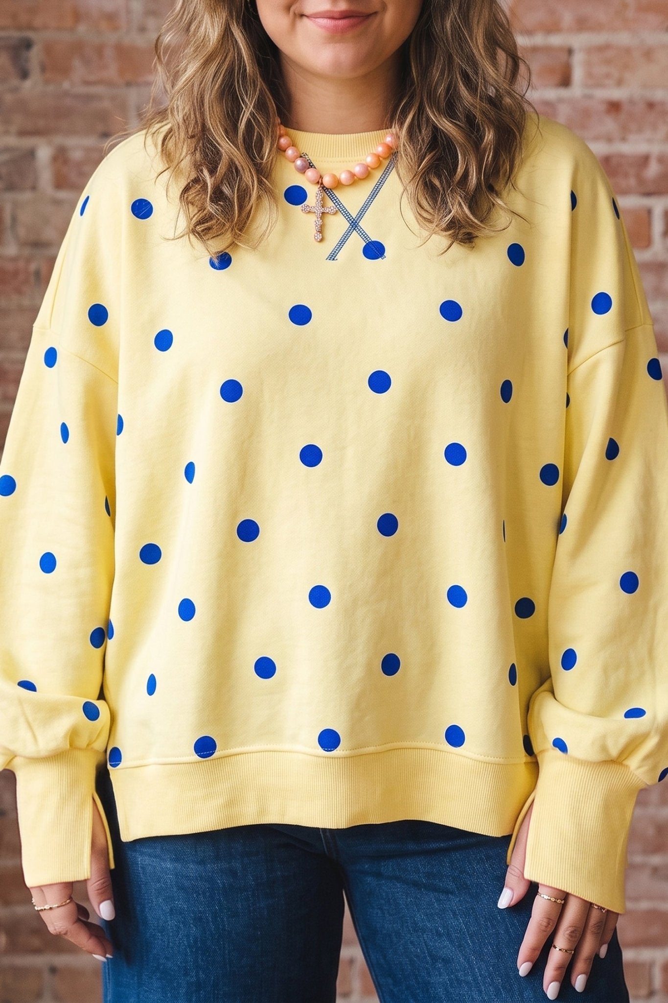 Woman in yellow blue-dot crewneck, blue jeans & beaded necklace by brick wall.