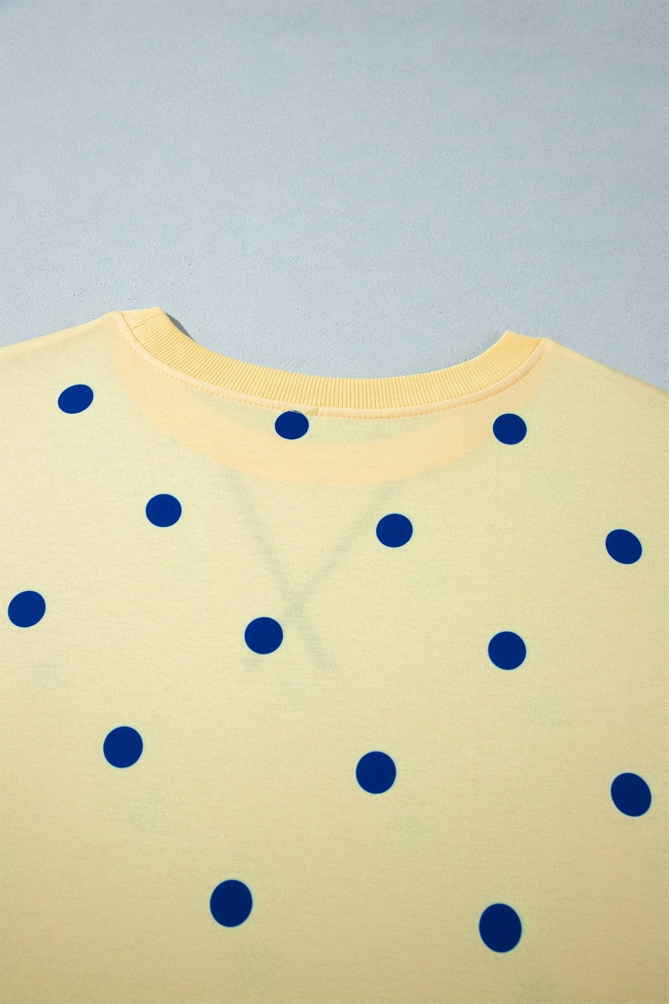 Close-up of pale yellow t-shirt with dark blue polka dots & ribbed collar on blue-gray.