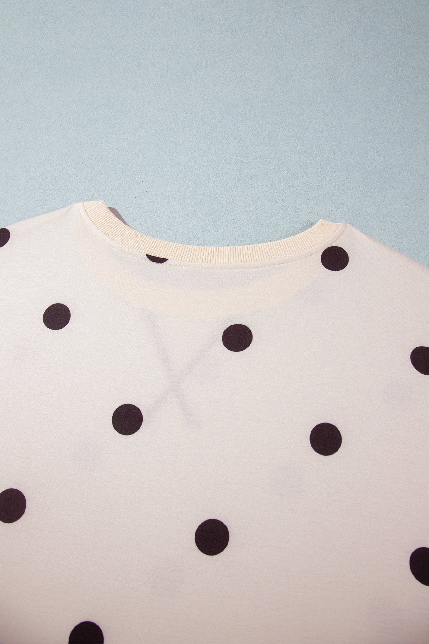 A close-up of a white crewneck top with black polka dots, shown against a light blue background.