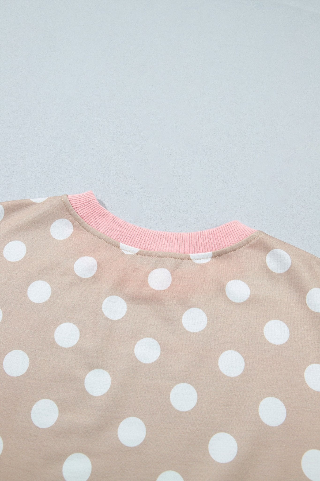 Close-up of beige polka-dot top with large white dots and pink ribbed crew neck on light gray.