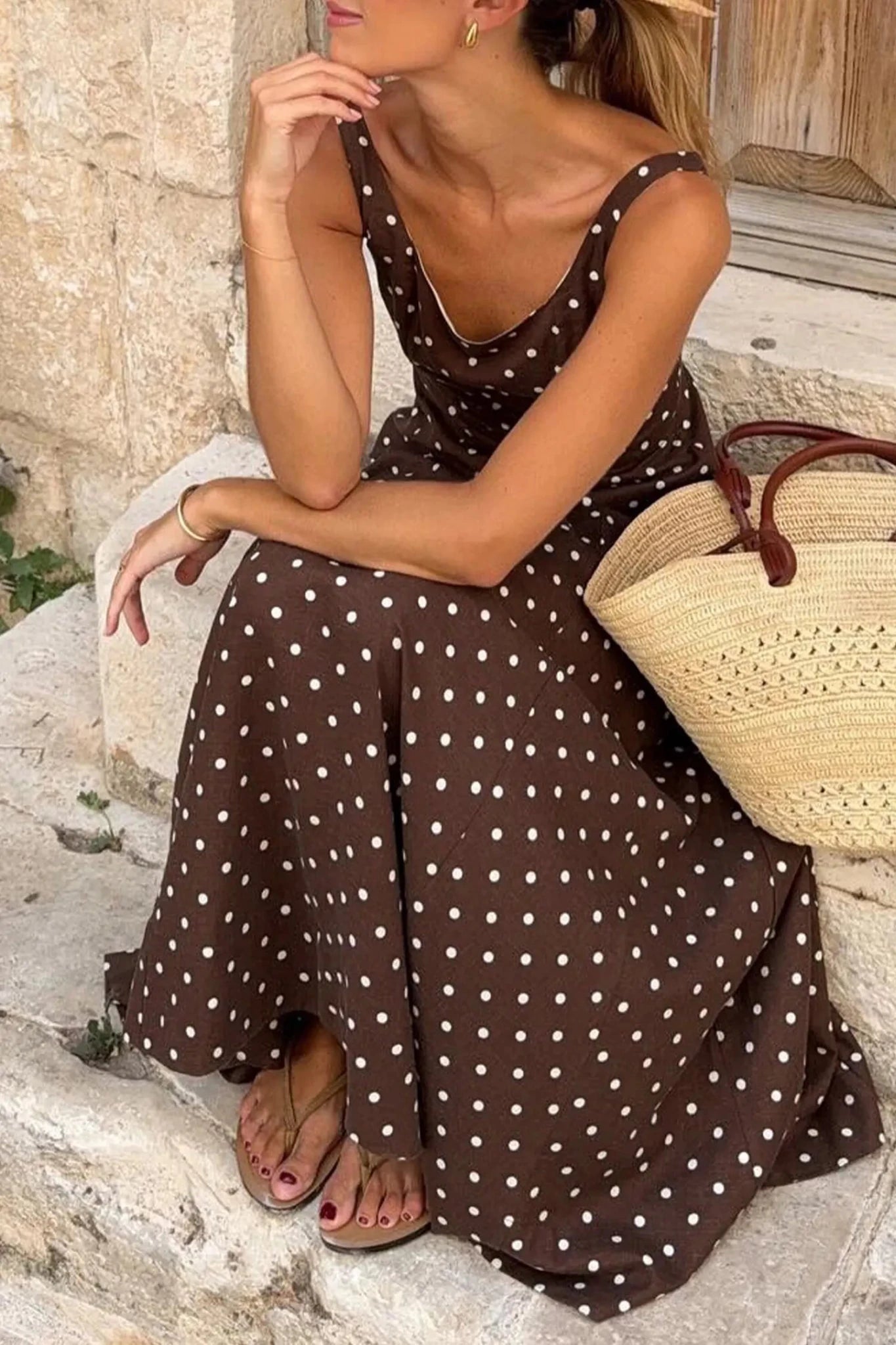 Woman in a brown polka dot dress sitting on stone steps with a straw bag.
