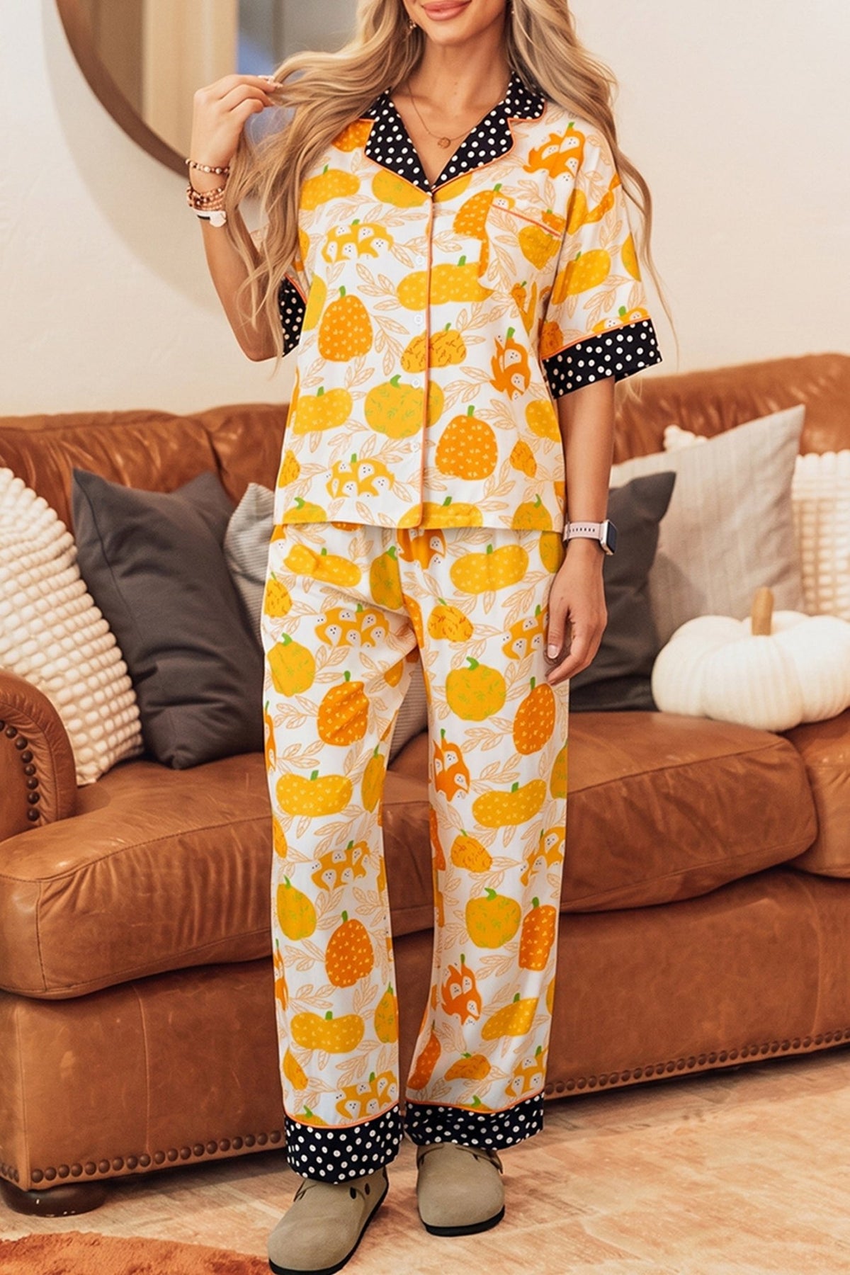 Woman in yellow and orange pumpkin-print pajamas by a brown leather couch.
