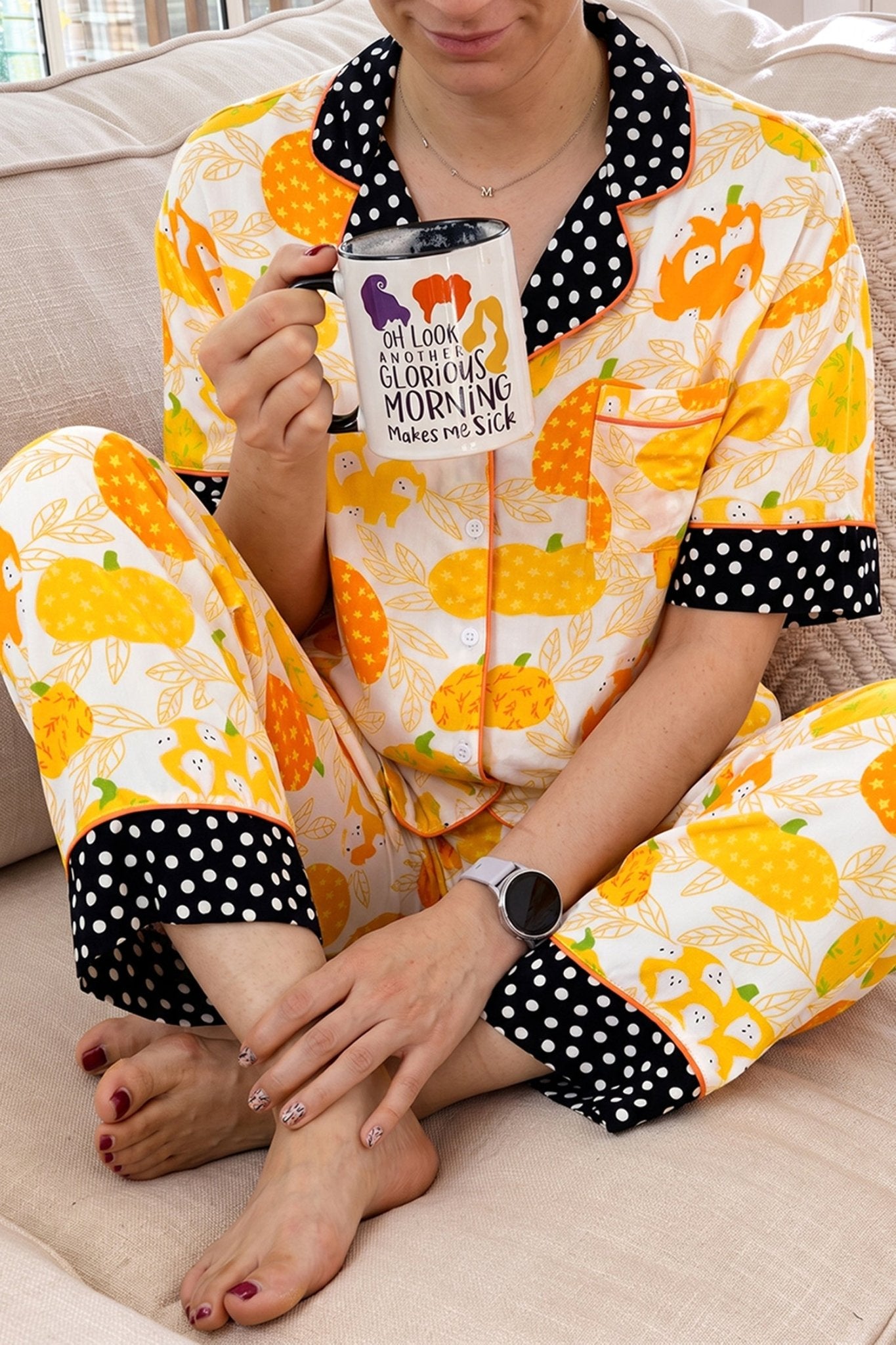 Person in pumpkin-print pajamas with polka-dot trim, holding a Halloween mug.

