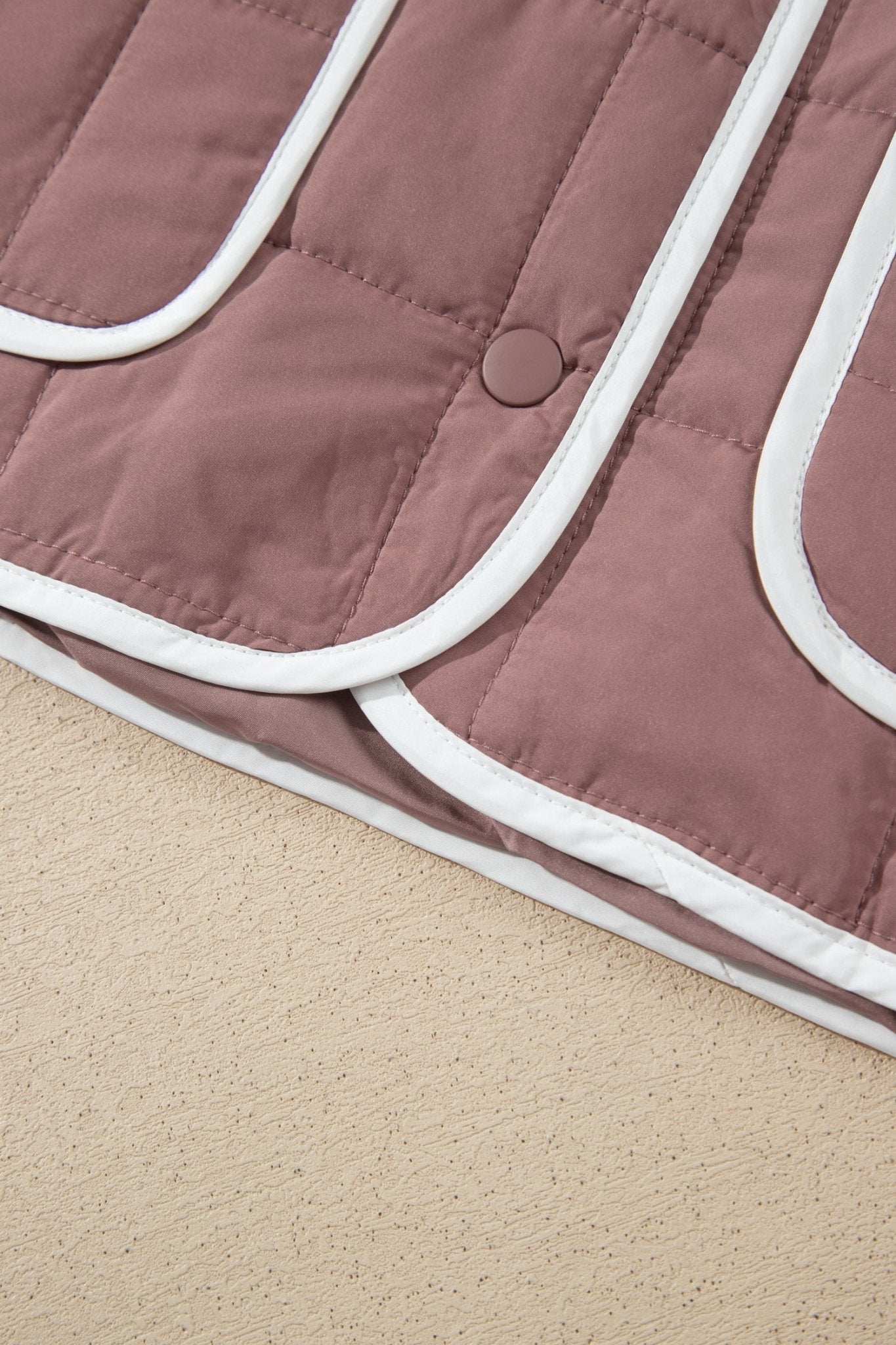 Close-up of dusty rose quilted jacket with white piping and snap closure on beige surface.
