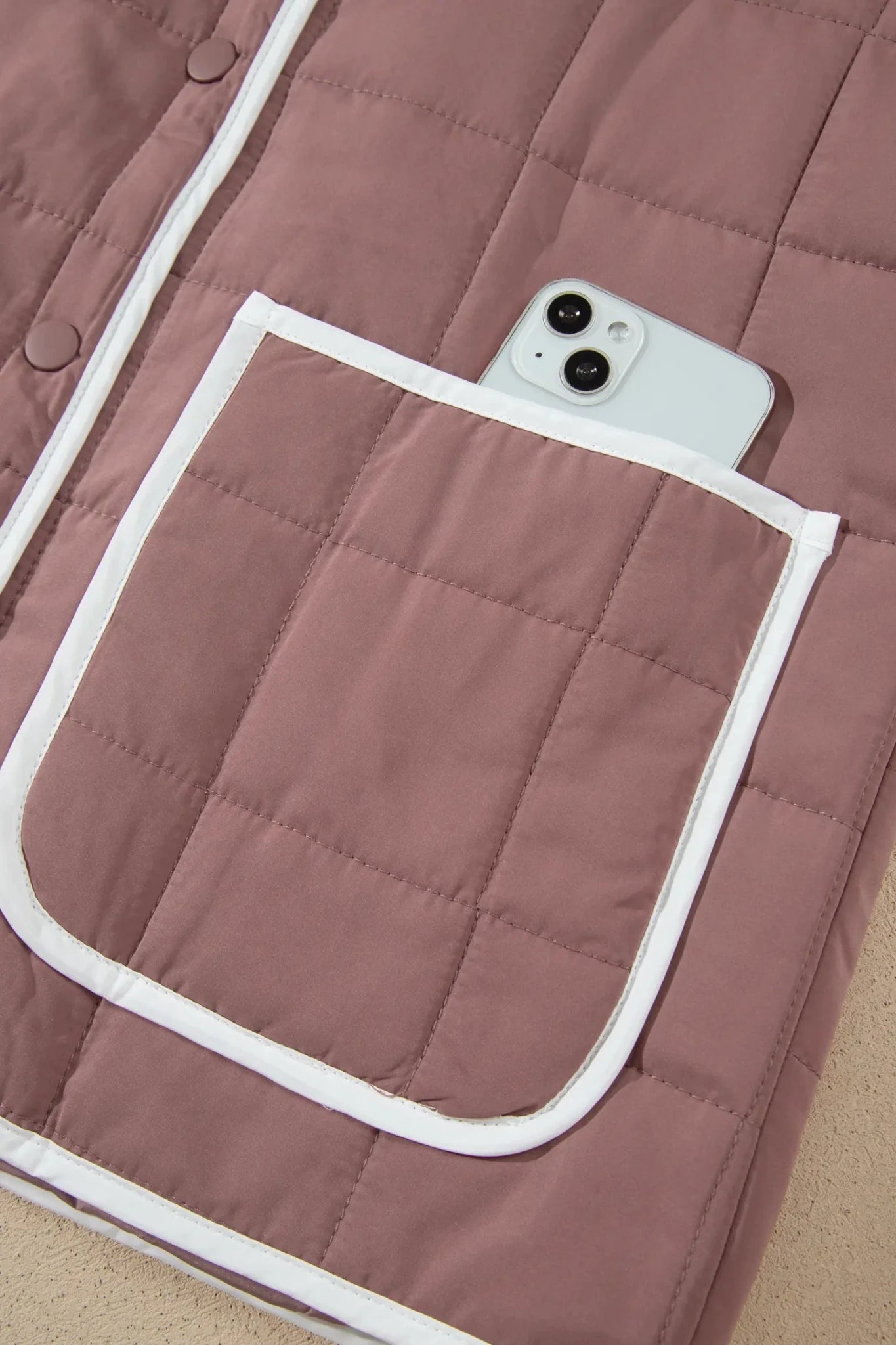 Close-up of dusty rose quilted jacket with white piping and large patch pocket holding a phone.