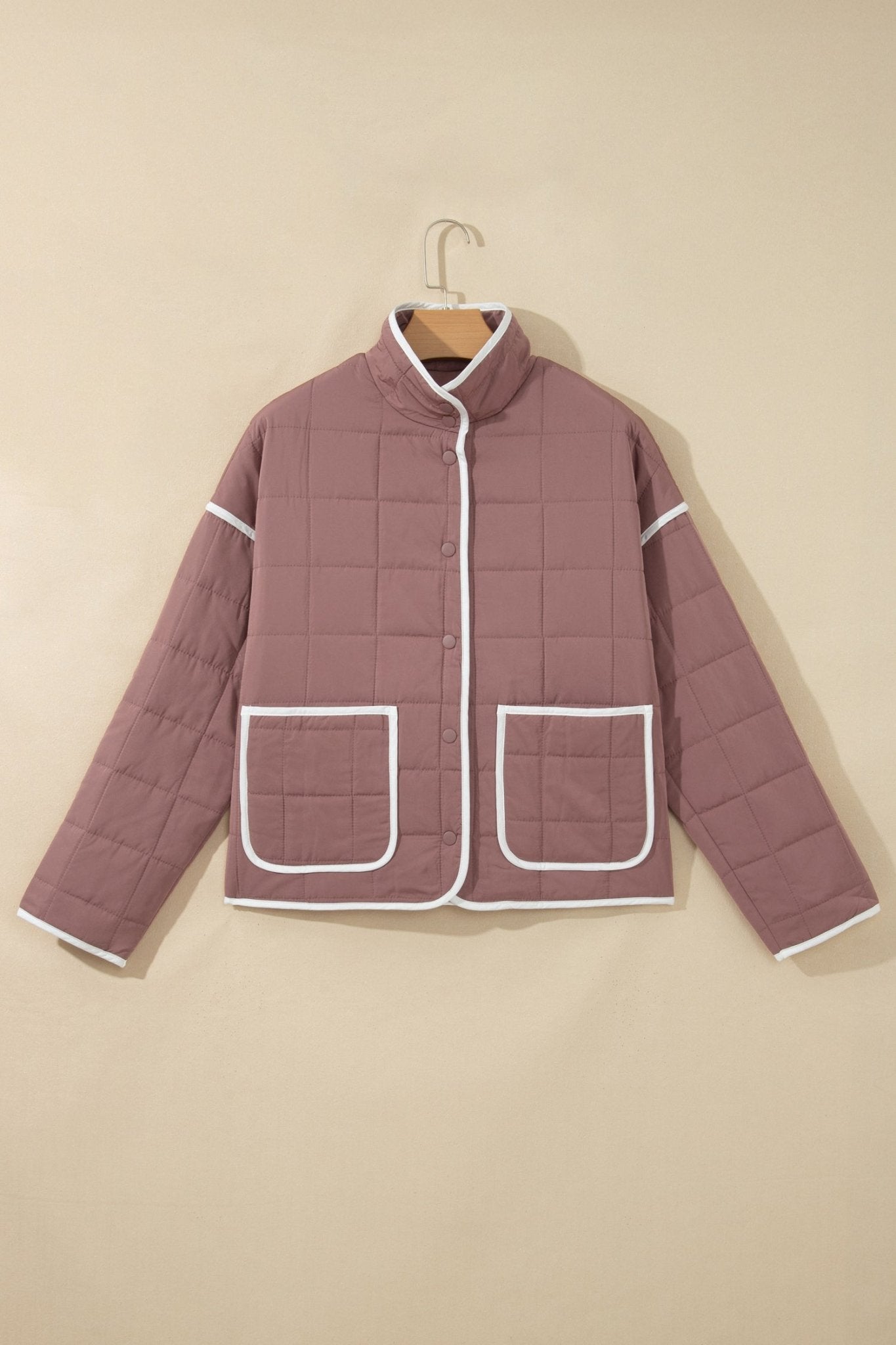 Dusty mauve quilted jacket with white piping trim hanging on a wooden hanger, beige background.