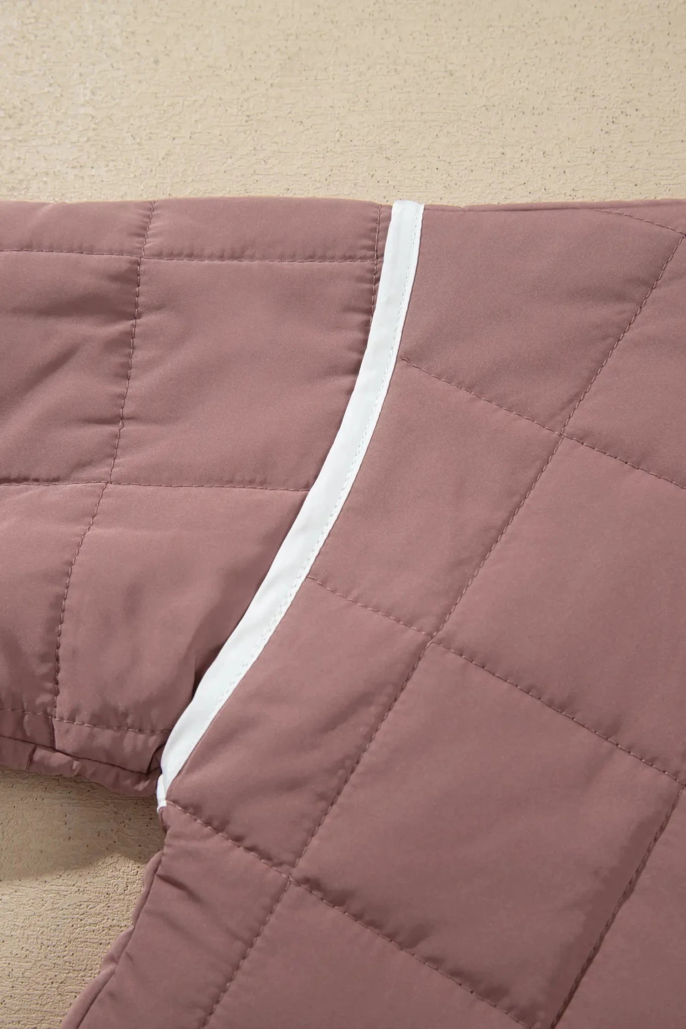 A close-up of a dusty rose quilted blanket with a white piping trim, laid flat on a beige carpet.