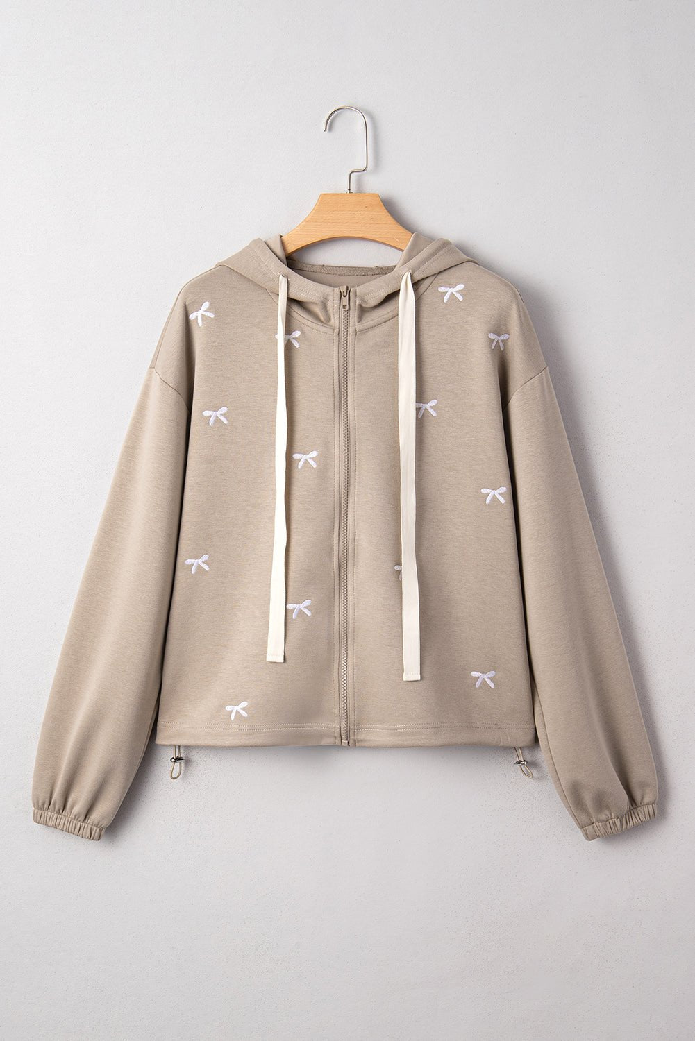 Beige zip-up hoodie with white drawstrings and embroidered bow motifs on a wooden hanger.