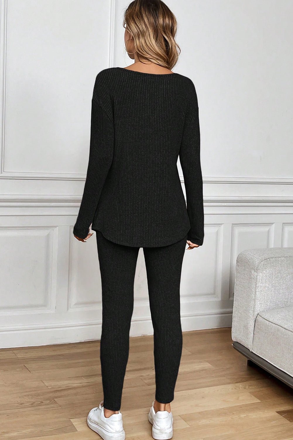 Person wearing a black ribbed sweater and leggings set in a room with white walls and wooden flooring.