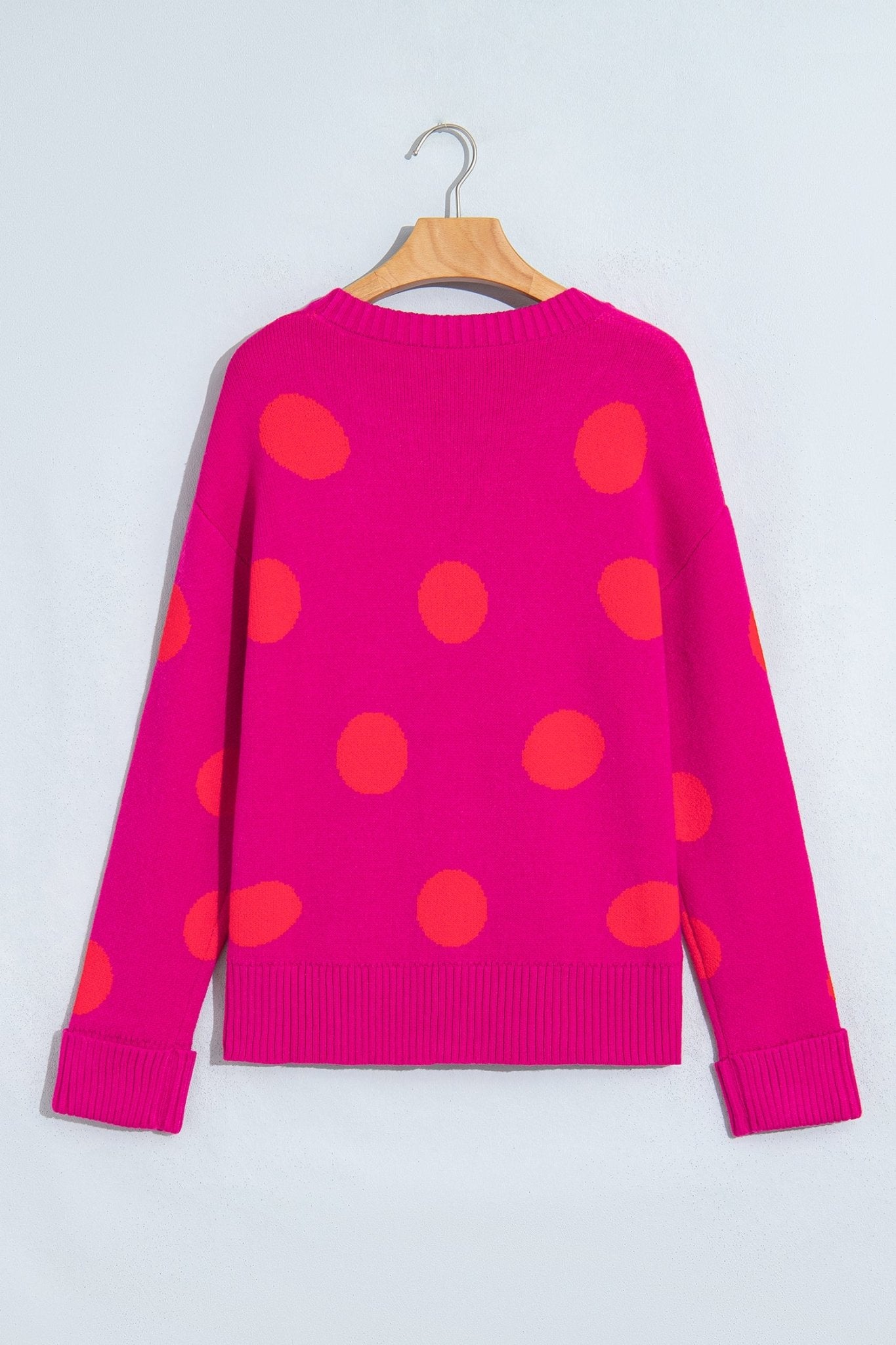 A bright pink knit sweater with large orange polka dots, hanging on a wooden hanger against a light gray wall.