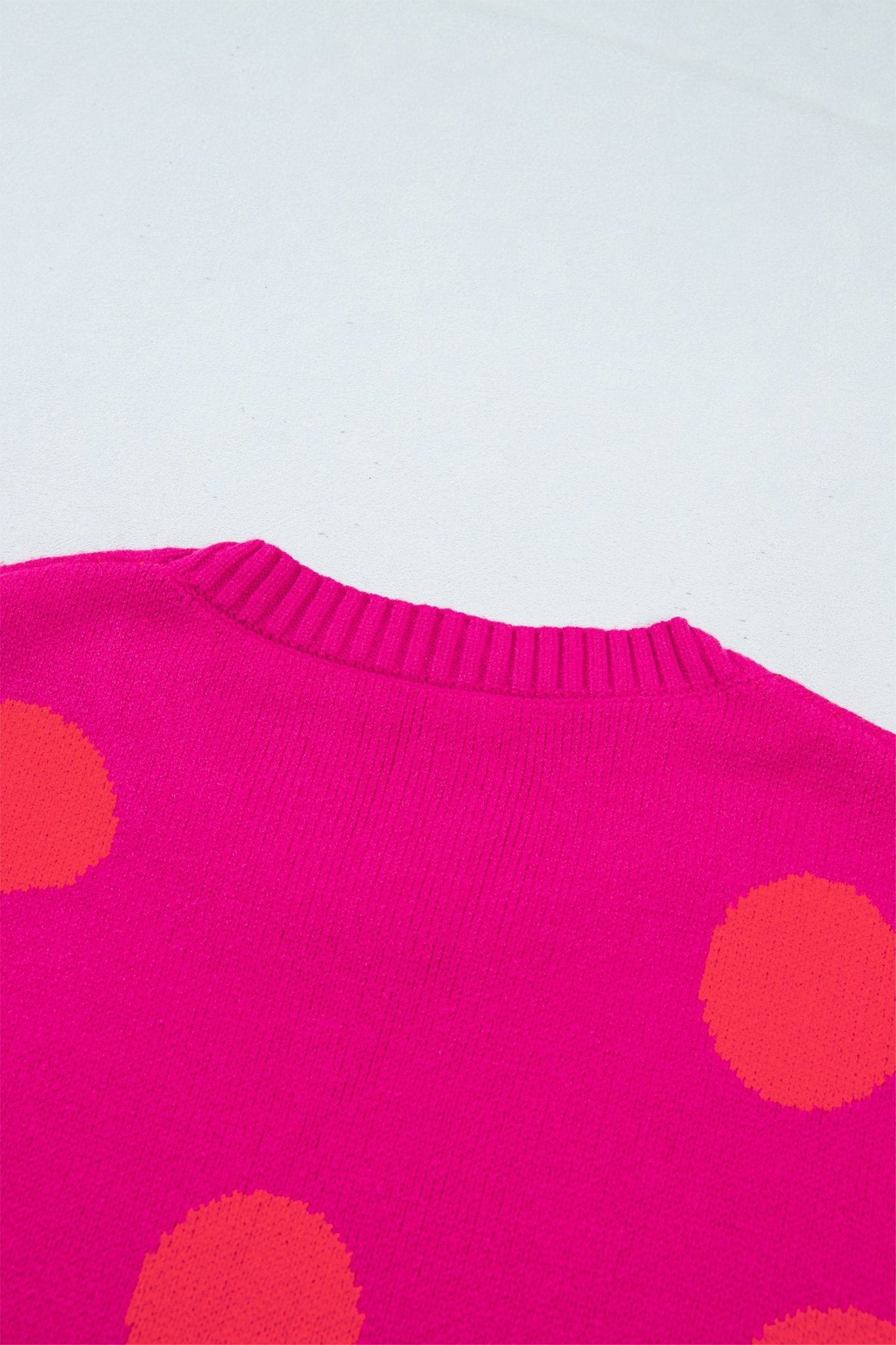 Close-up of bright pink polka-dot knit sweater with ribbed crew neck on plain light background.