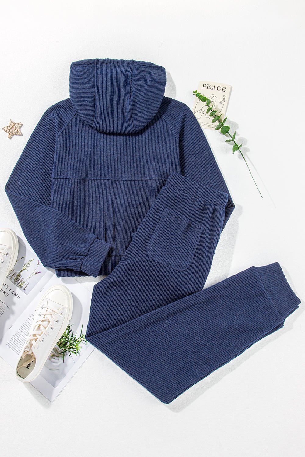 Navy blue hoodie and pants set on a white background with a plant and magazine.