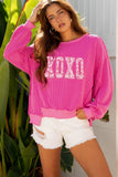 Woman wearing a bright pink sweater with text and white shorts, standing outdoors.