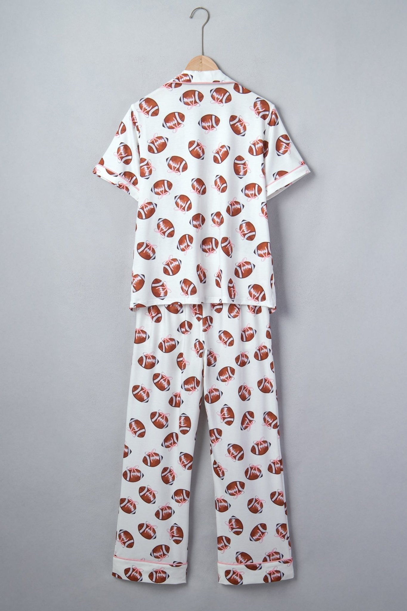 A two-piece short-sleeve pajama set hanging on a wooden hanger against a plain gray background. The pajama top and matching pants are white with an all-over repeating pattern of brown and white footballs.
