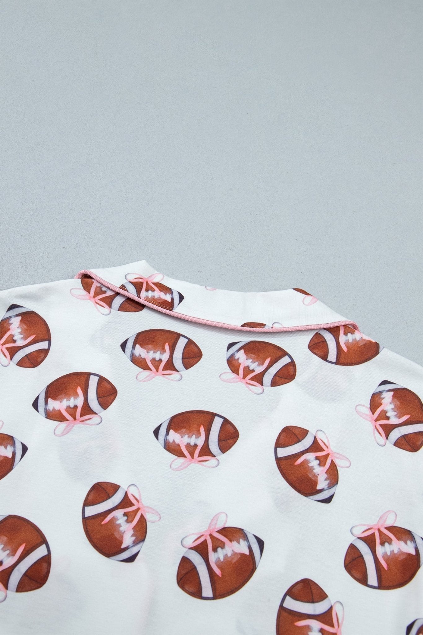 A close-up of a white garment featuring an all-over print of brown footballs with pink bows, set against a solid light gray background.