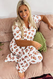 A woman sitting on a dusty pink couch wearing a white short-sleeve pajama set with an all-over brown football print. She is holding a green fringed throw pillow.