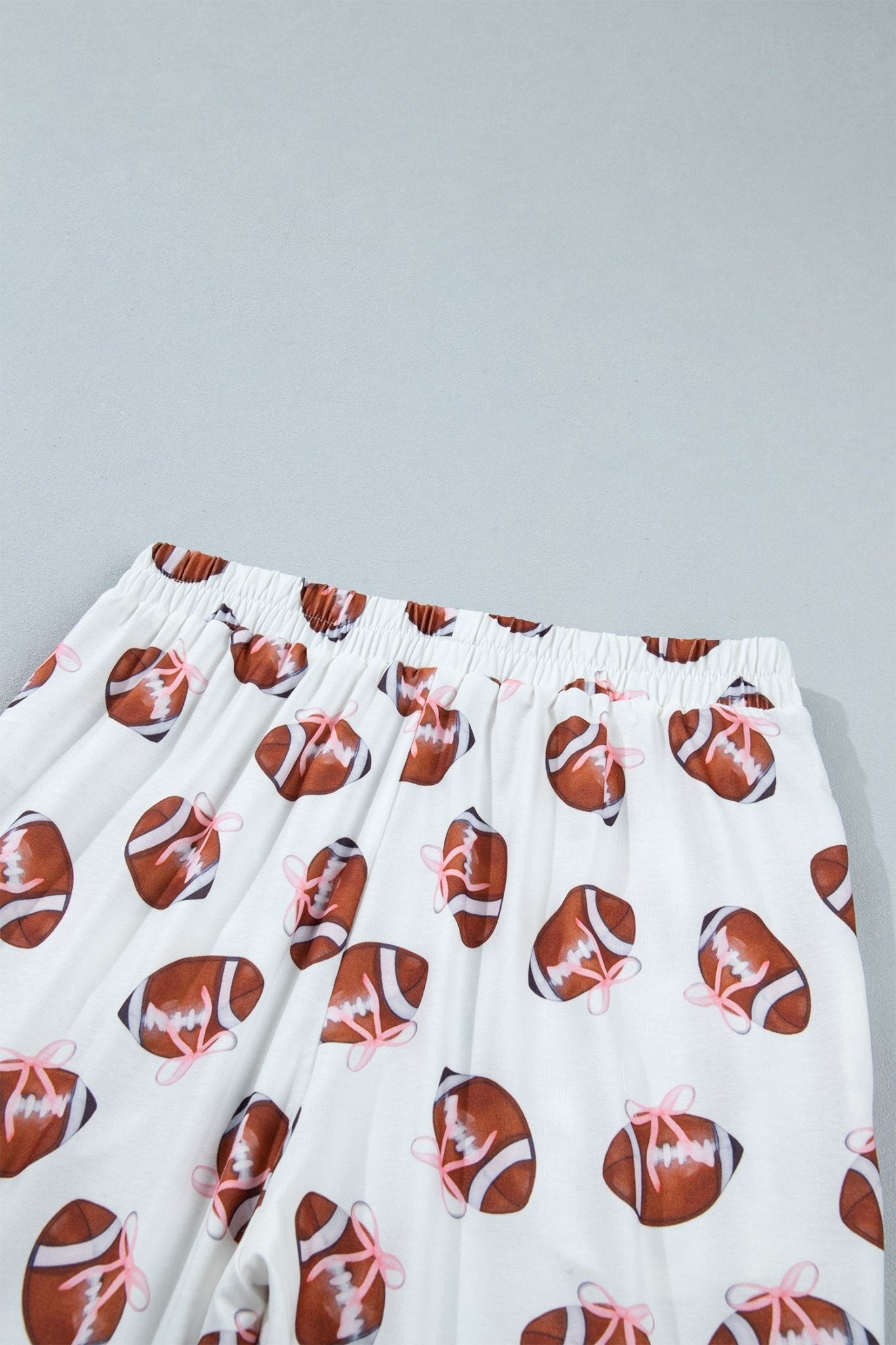 White lounge pants with an all-over print of brown footballs decorated with pink bows, shown against a light gray background.