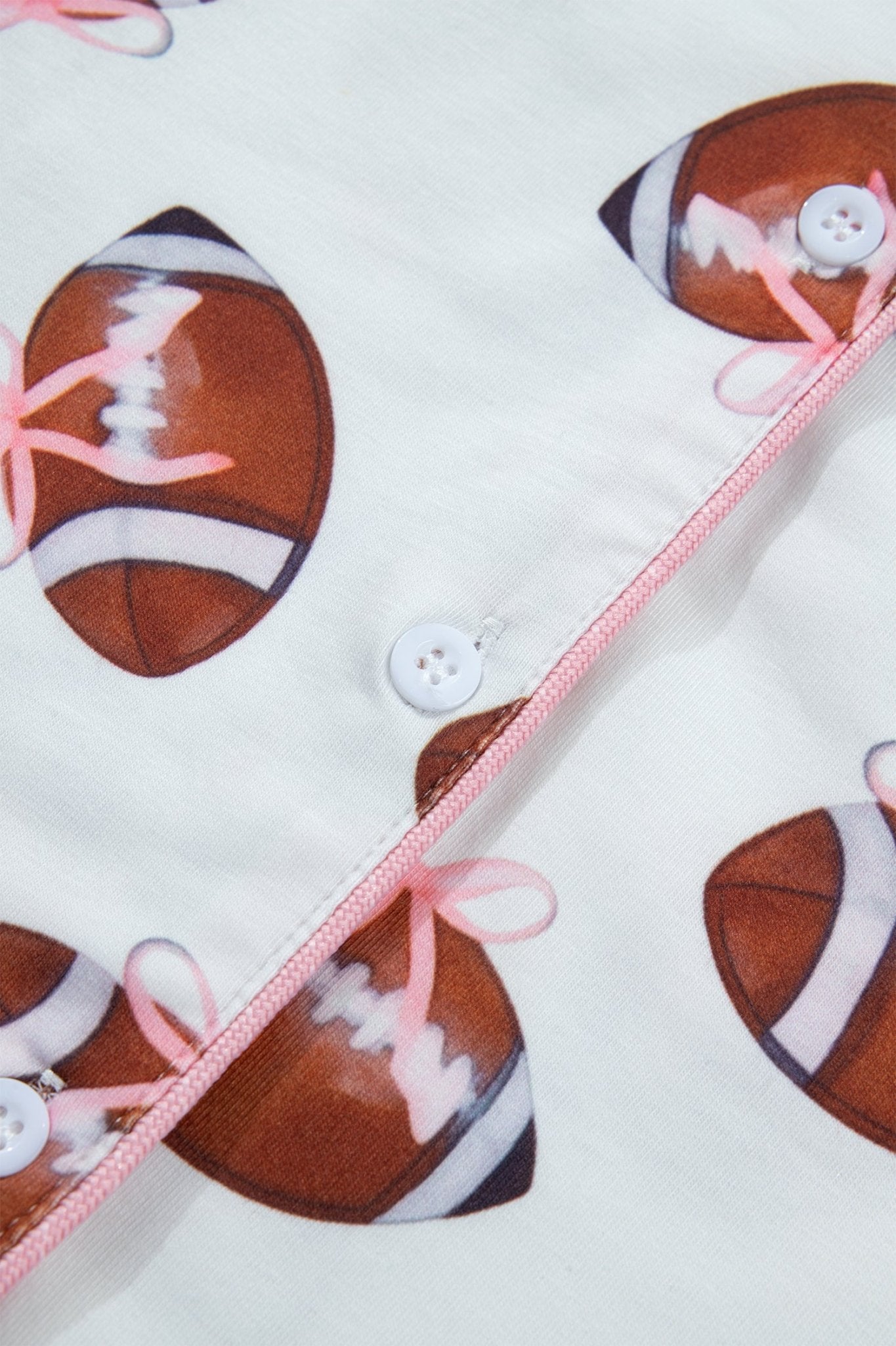 Close-up of a white fabric product featuring a repeating brown football print with pink laces, pink trim, and white buttons.