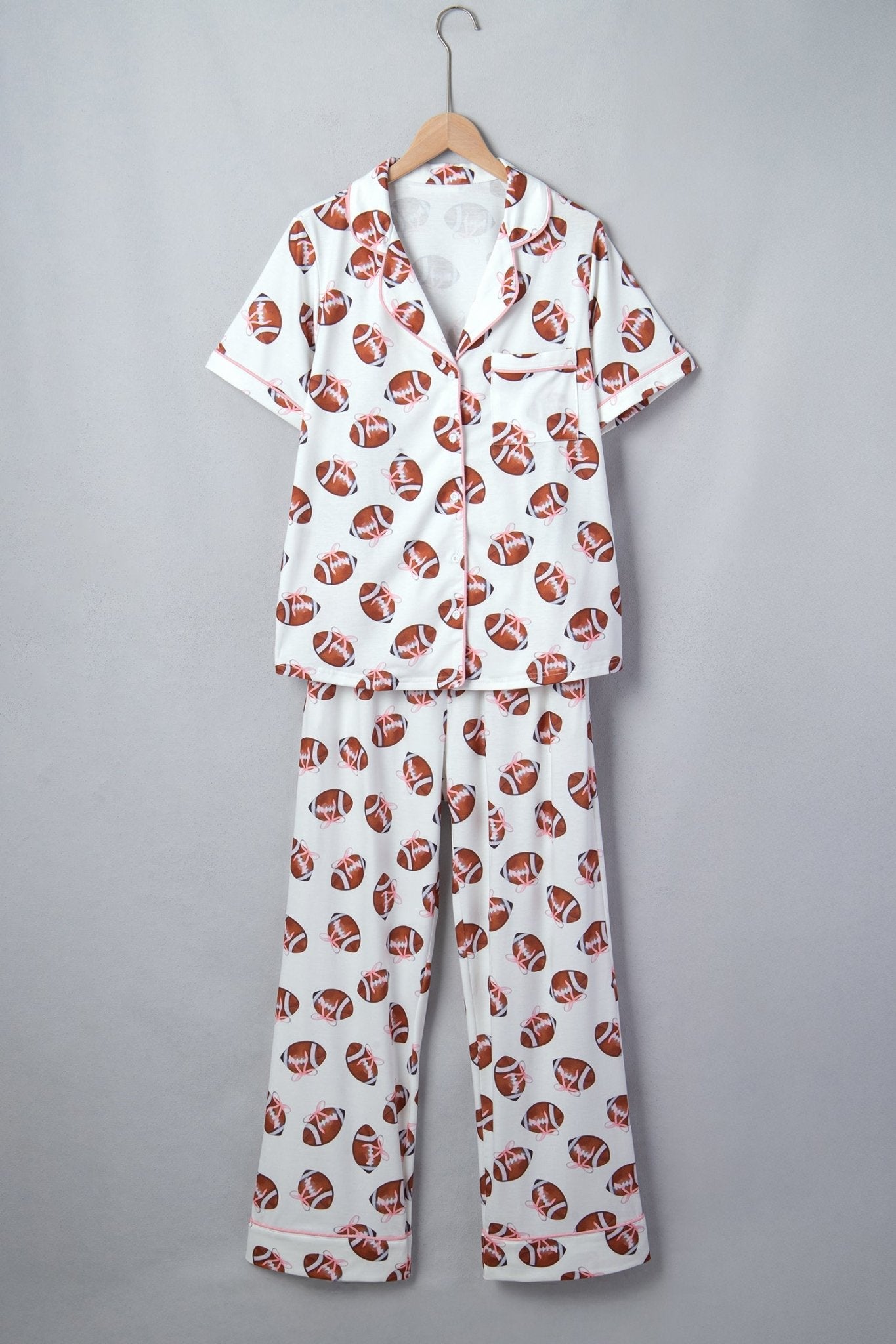 A two-piece short-sleeve pajama set hanging on a wooden hanger against a plain gray background. The pajama set features a white base fabric with an all-over print of brown and white footballs.