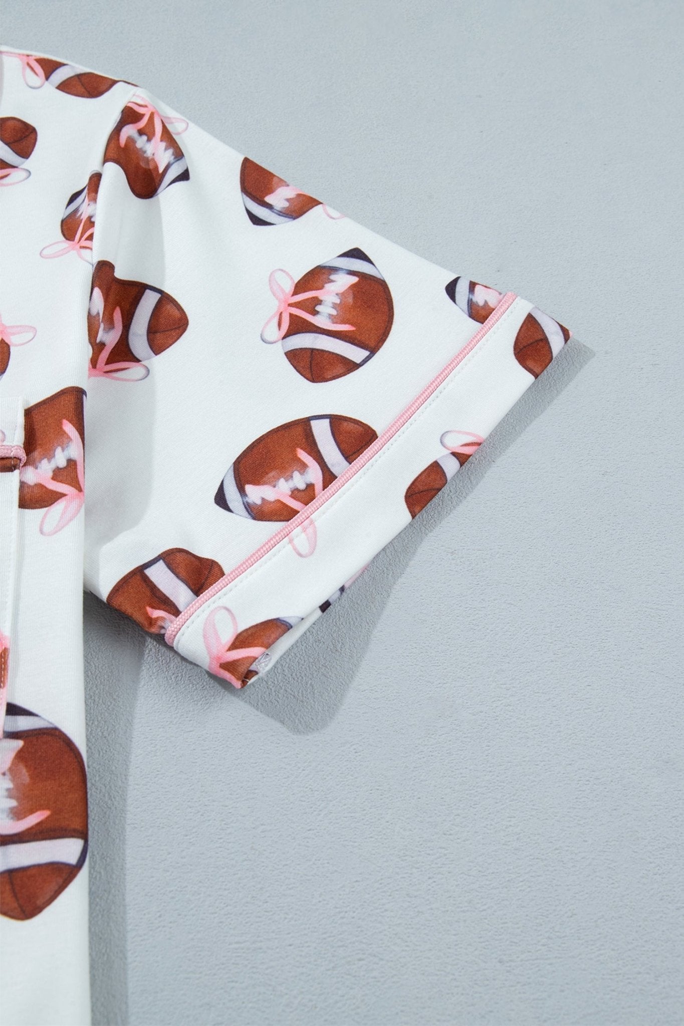 A close-up of a white pajama or robe sleeve featuring an all-over print of brown footballs with pink laces, finished with a light pink piped cuff, set against a neutral gray background.