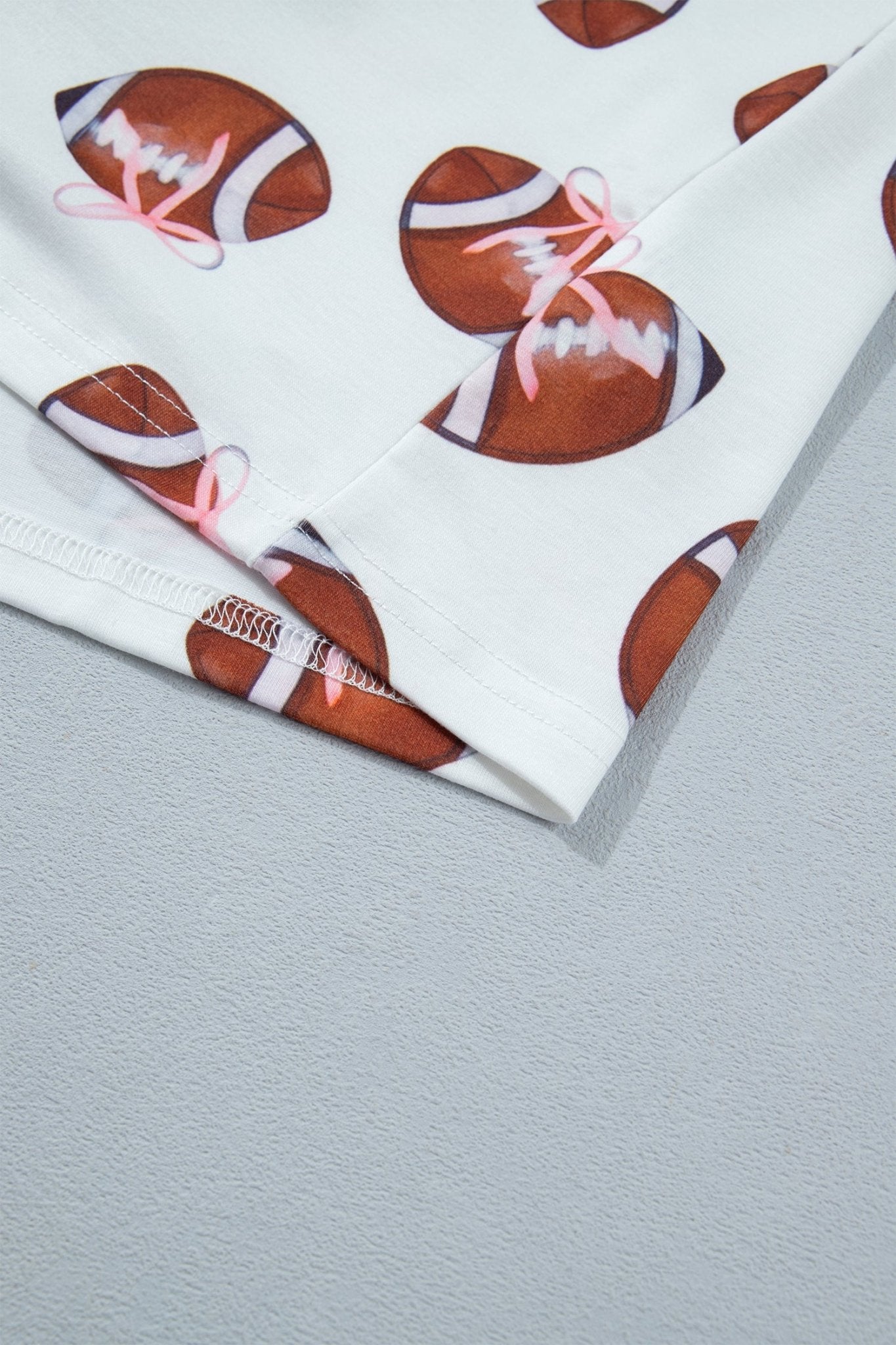 A close-up of a white fabric product, likely a towel or blanket, featuring a repeating pattern of brown American footballs with white stripes and pink bows, laid flat on a light gray textured surface.